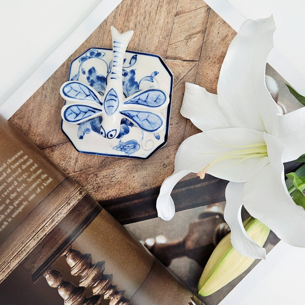 A small octagonal ceramic lid with a blue and white dragonfly design, styled on a wooden surface next to a white lily flower and an open book.