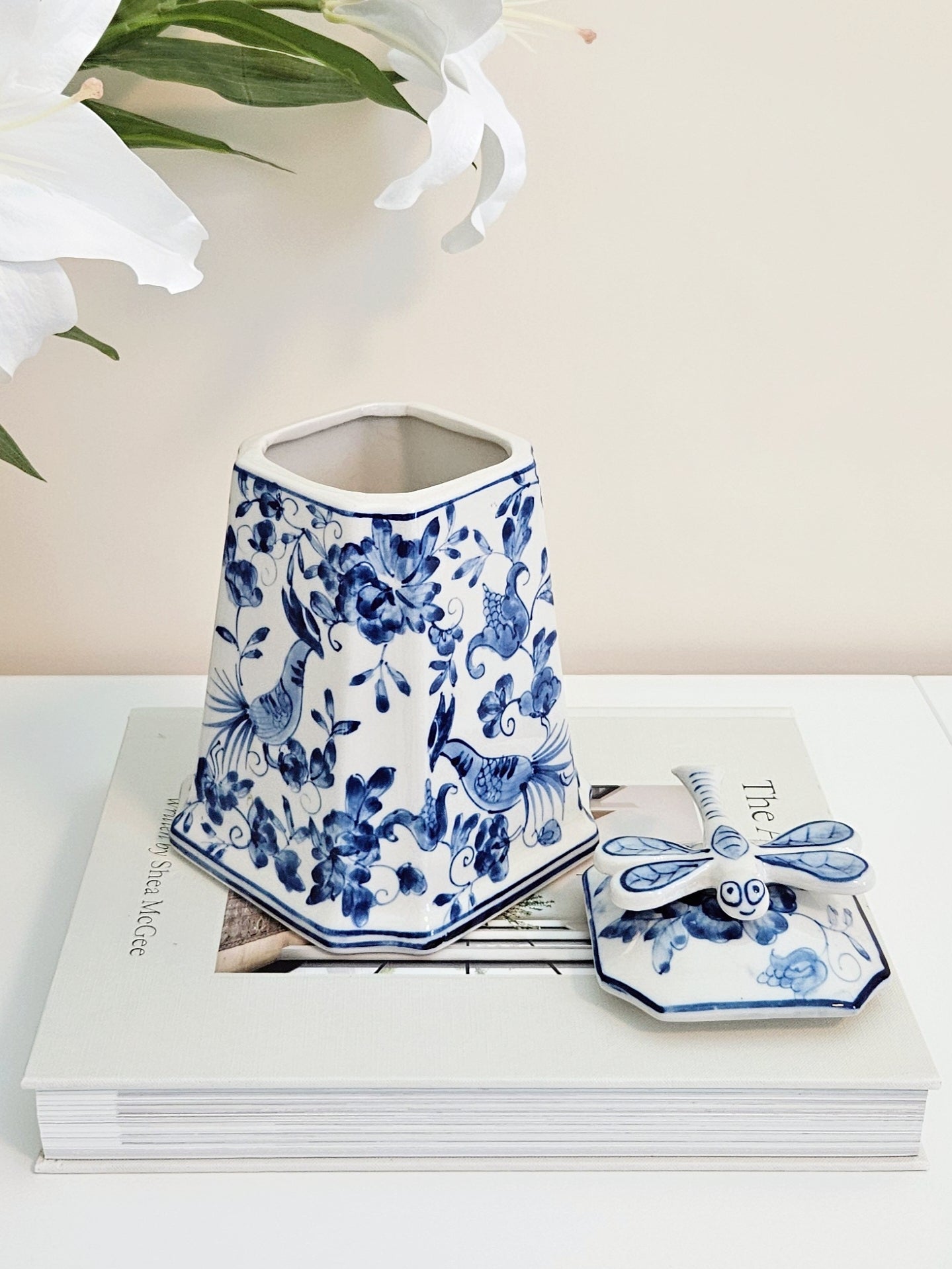 Blue and white porcelain decorative canister with a matching dragonfly lid, displayed on a stack of books against a light beige background with white flowers in the corner.