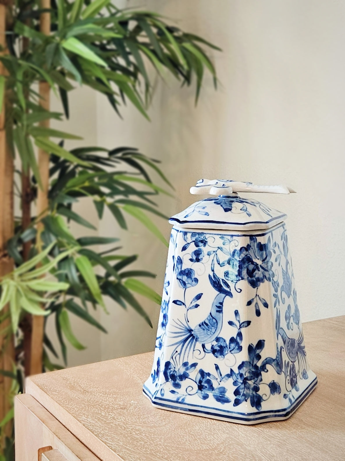 A chinoiserie blue and white porcelain  jar with a lid, decorated with traditional Chinese bird and floral motifs, sitting on a light wood table next to a green plant.