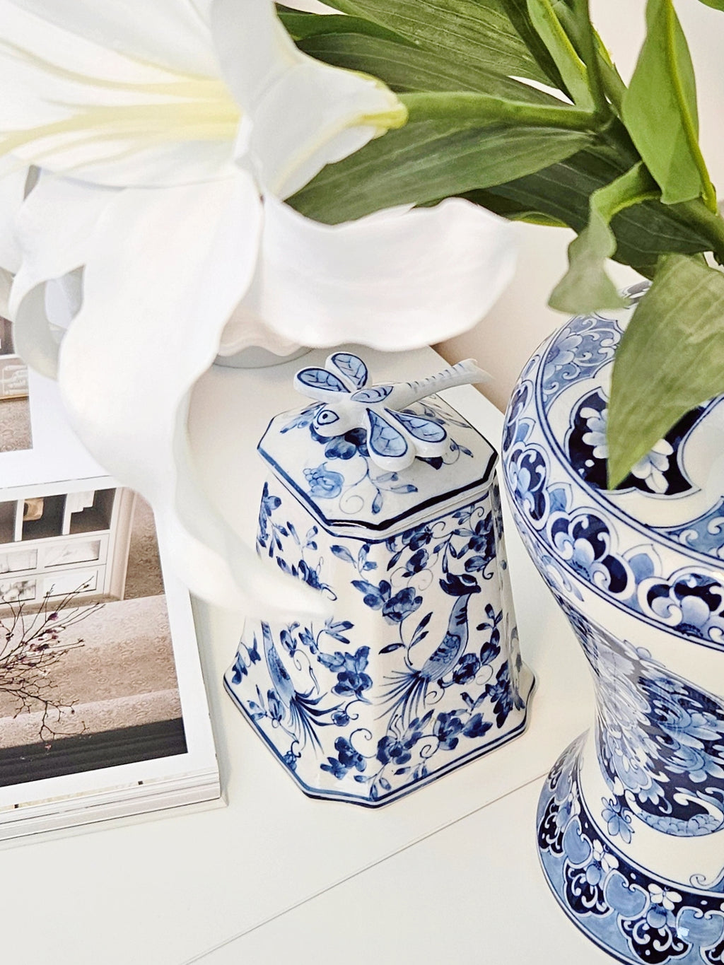 A blue and white porcelain lidded jar with floral patterns sits on a white surface next to a matching blue and white vase, with a large white lily flower and green leaves in the foreground.