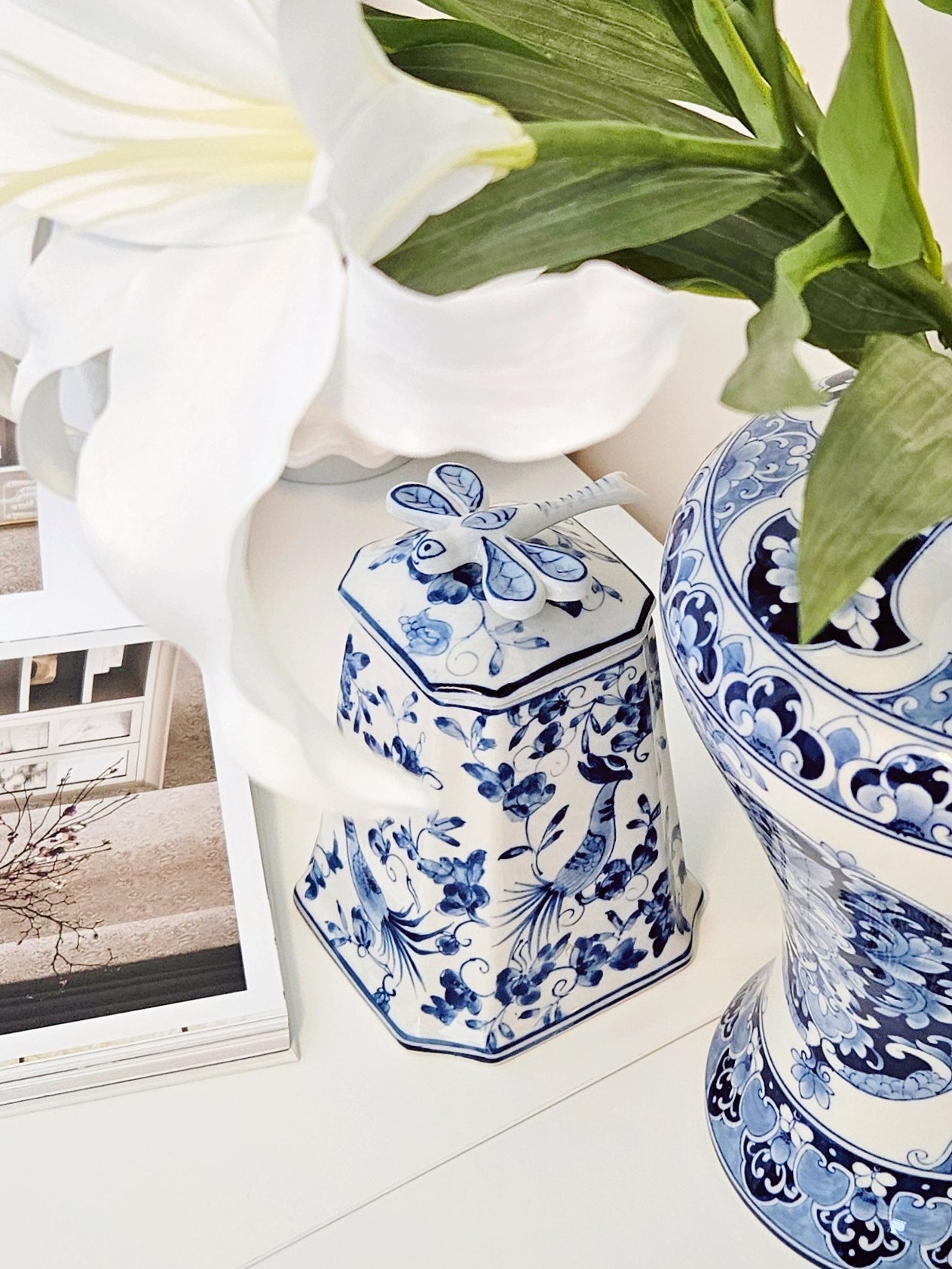 A blue and white porcelain lidded jar with floral patterns sits on a white surface next to a matching blue and white vase, with a large white lily flower and green leaves in the foreground.