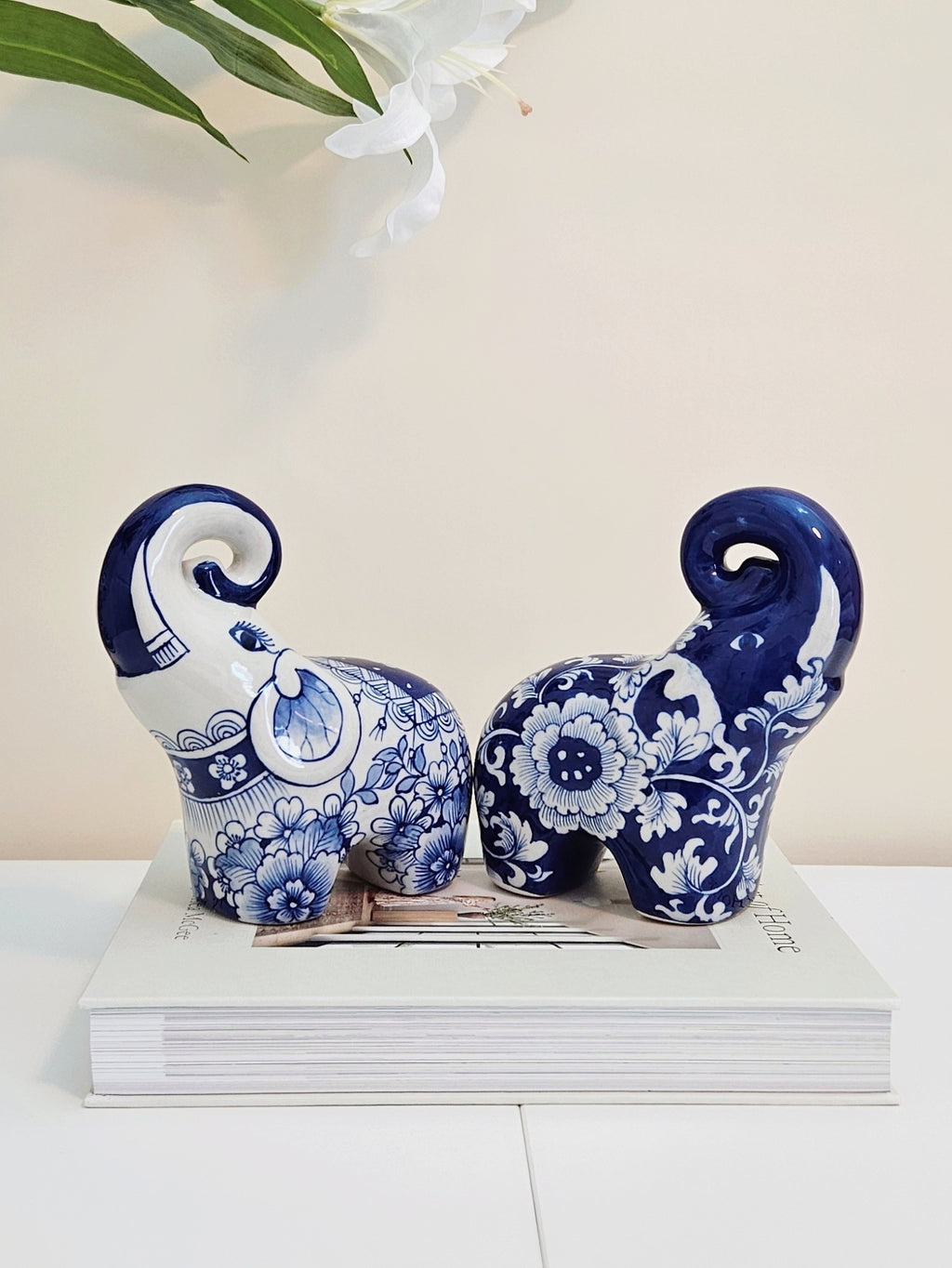 Two chinoiserie decorative ceramic elephants in a blue and white floral pattern, displayed on top of a white book against a light beige background.