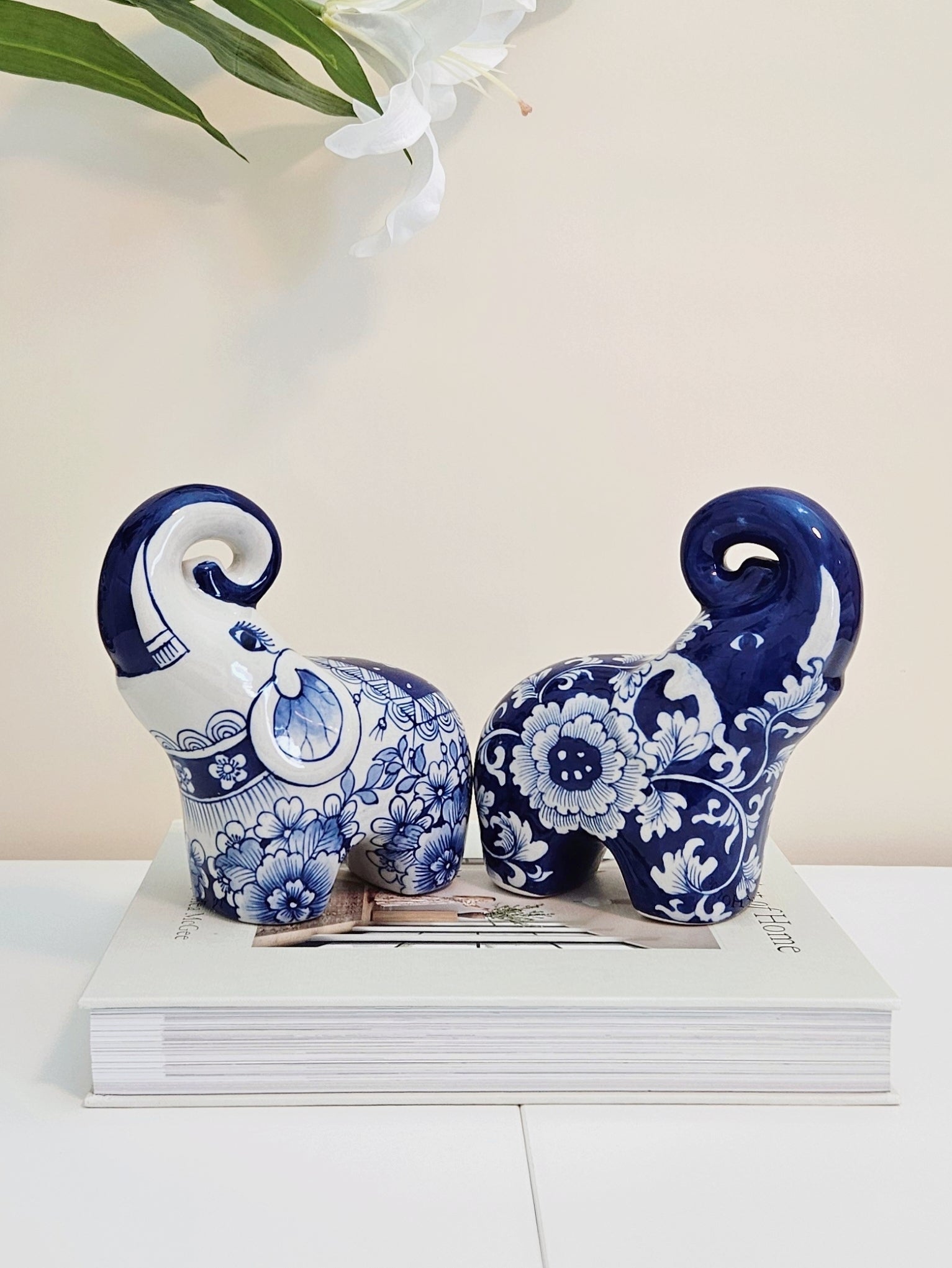 Two chinoiserie decorative ceramic elephants in a blue and white floral pattern, displayed on top of a white book against a light beige background.