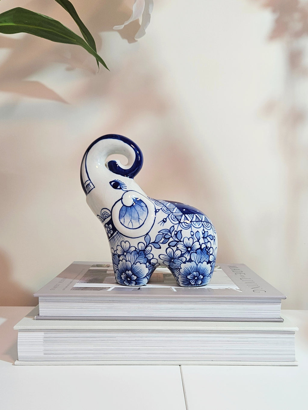 A chinoiserie blue and white ceramic elephant figurine with floral patterns, displayed on top of two stacked books against a soft beige background with a leaf in the upper left corner.
