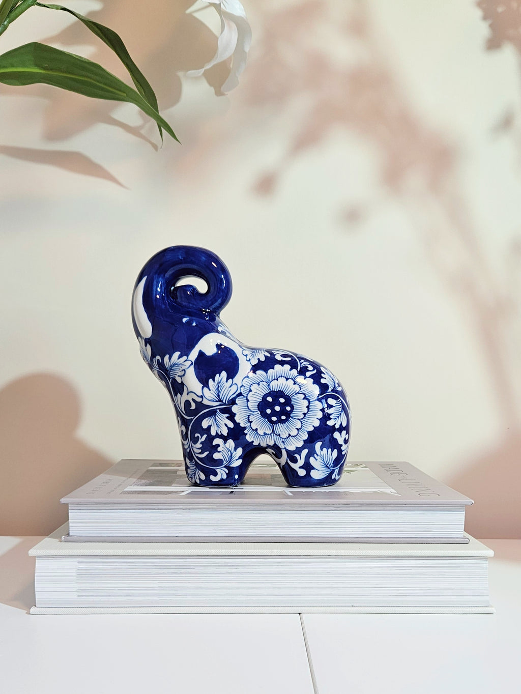 A glossy blue and white porcelain elephant figurine with floral patterns, displayed on a stack of white books against a softly lit beige background with leaf shadows.