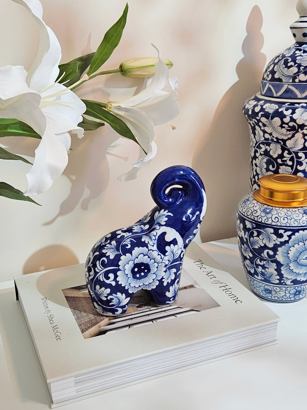 A small glossy chinoiserie blue and white ceramic elephant figurine with floral patterns sits on top of a white book. To the right is a matching blue-and-white decorative jar with a gold lid, and white lilies are visible in the background against a warm beige wall.