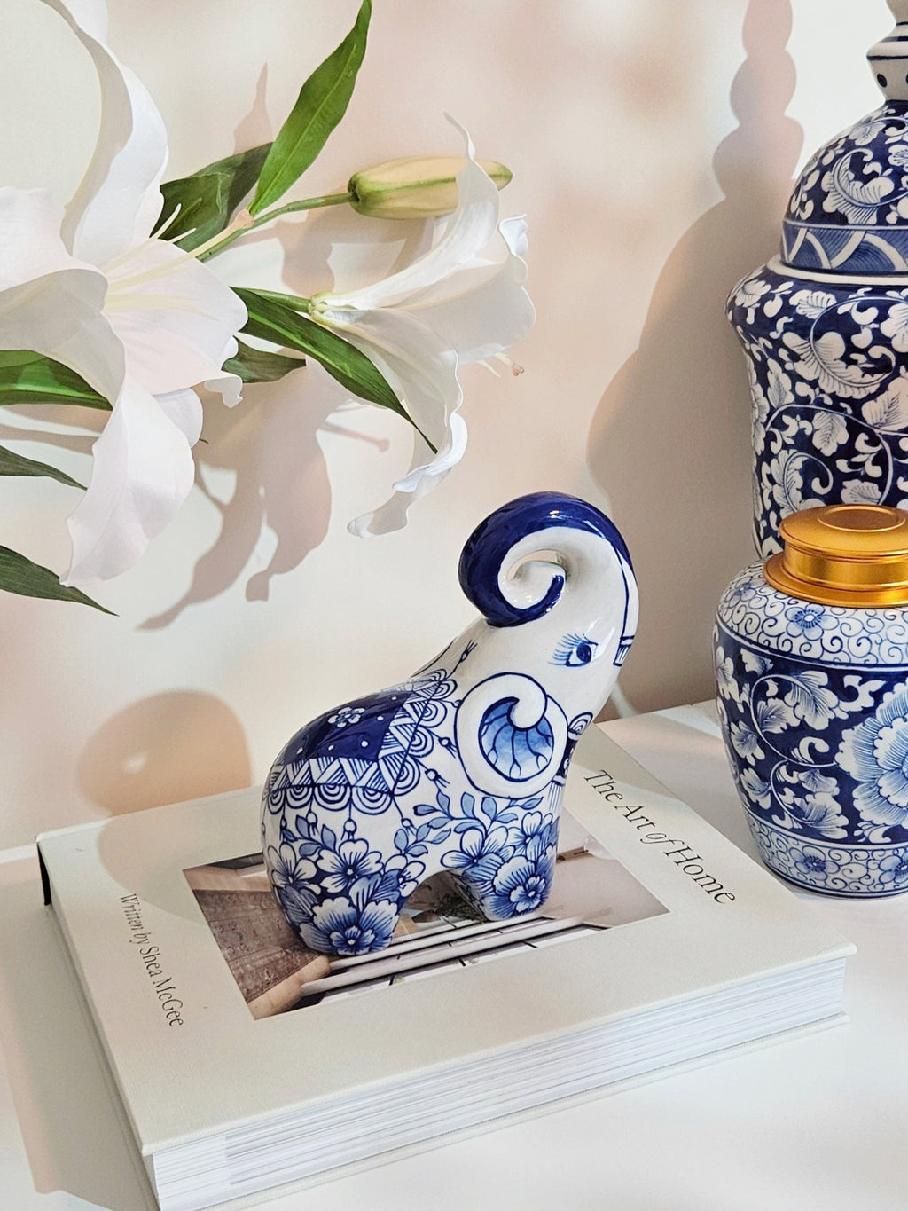A blue and white porcelain elephant figurine with floral patterns sits on a white book, styled with white lilies and blue-and-white ceramic vases against a warm beige background.