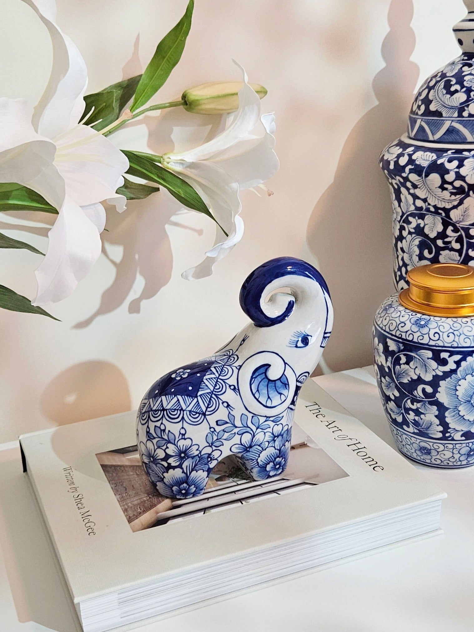 A blue and white porcelain elephant figurine with floral patterns sits on a white book, styled with white lilies and blue-and-white ceramic vases against a warm beige background.
