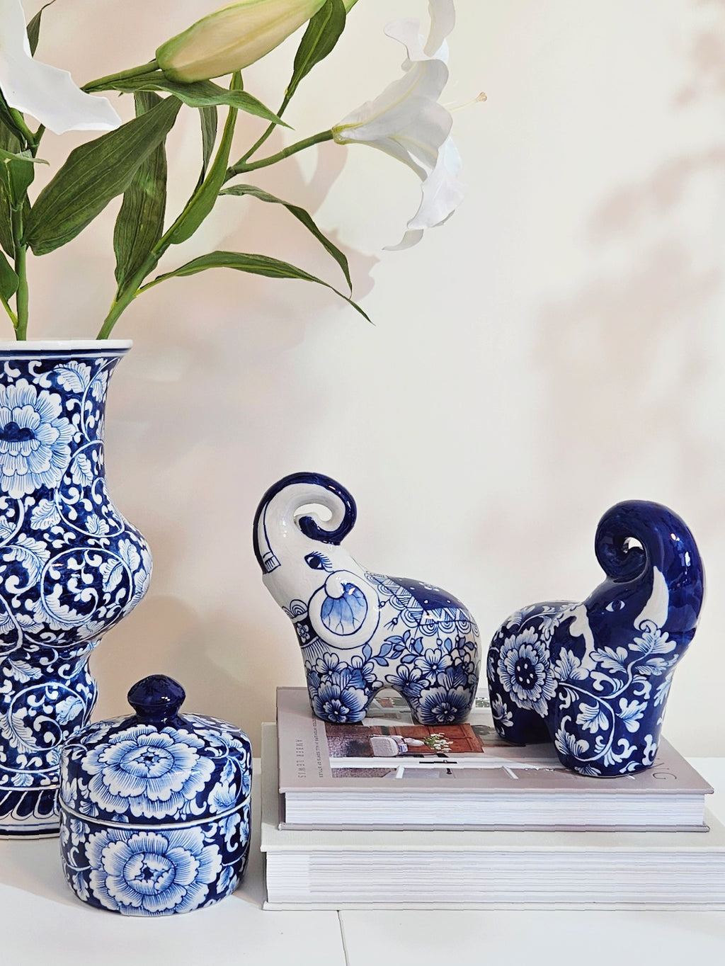 Blue and white floral ceramic elephant figurines displayed on books next to a matching blue and white floral vase with white lilies on a white surface.
