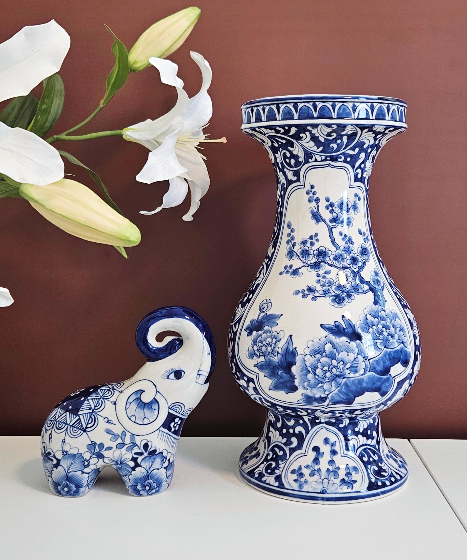 A blue and white porcelain vase with floral patterns stands on a white surface against a dark brown wall, next to a matching blue and white chinoiserie elephant figurine. White lily flowers are draped on the left side.