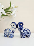 Two chinoiserie decorative porcelain elephants in a blue and white floral pattern, displayed on a white surface with white lilies in the background.