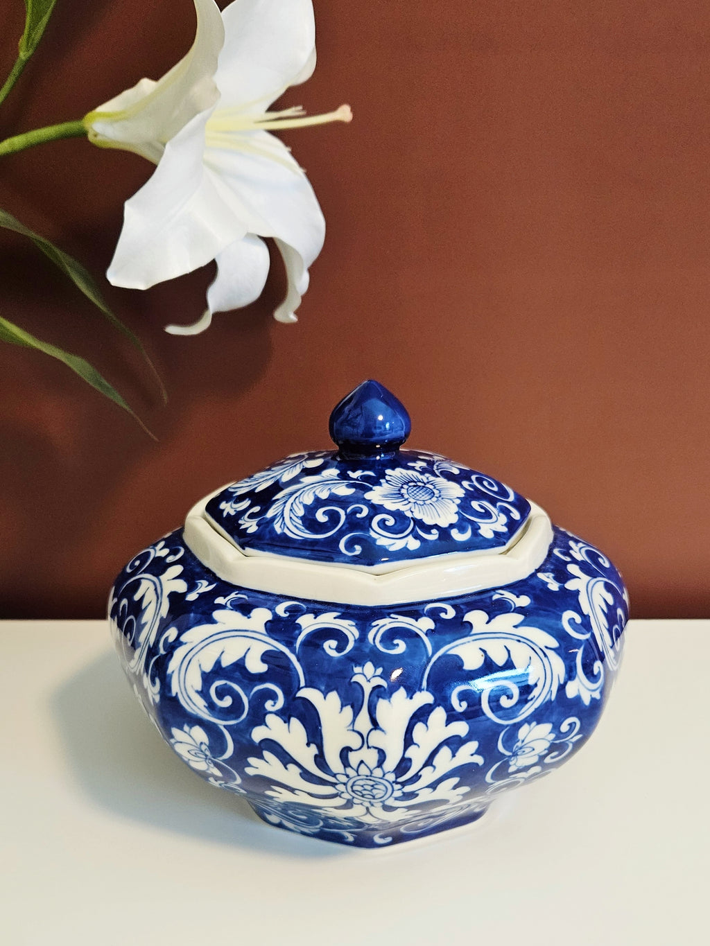 A chinoiserie  blue and white porcelain lidded jar with ornate floral scrollwork, placed on a white surface against a warm brown background, with a white lily flower in the upper left.