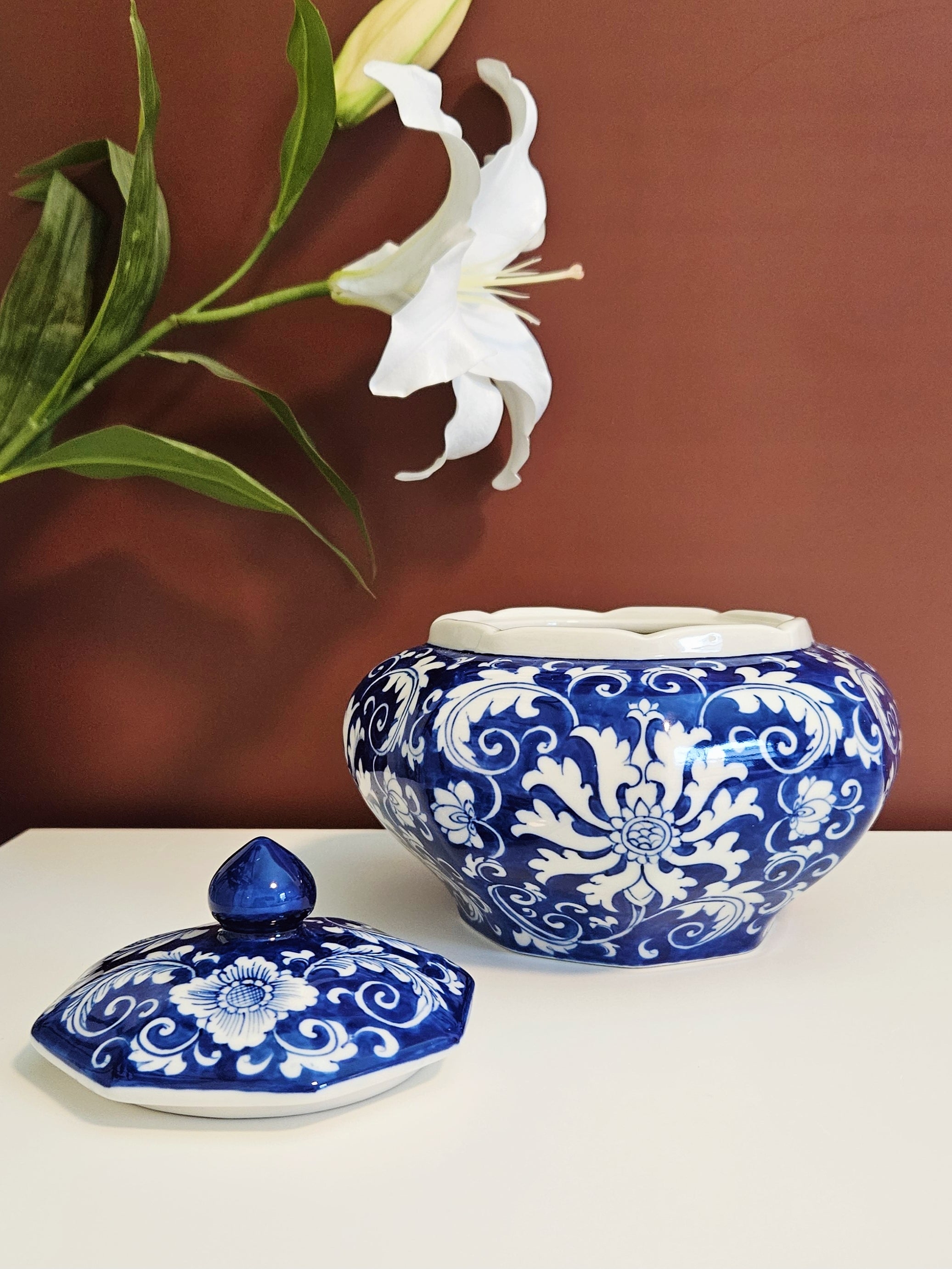 A blue and white porcelain lidded jar with ornate floral scrollwork, shown alongside its matching lid on a white surface against a warm brown wall, with a white lily flower in the background.