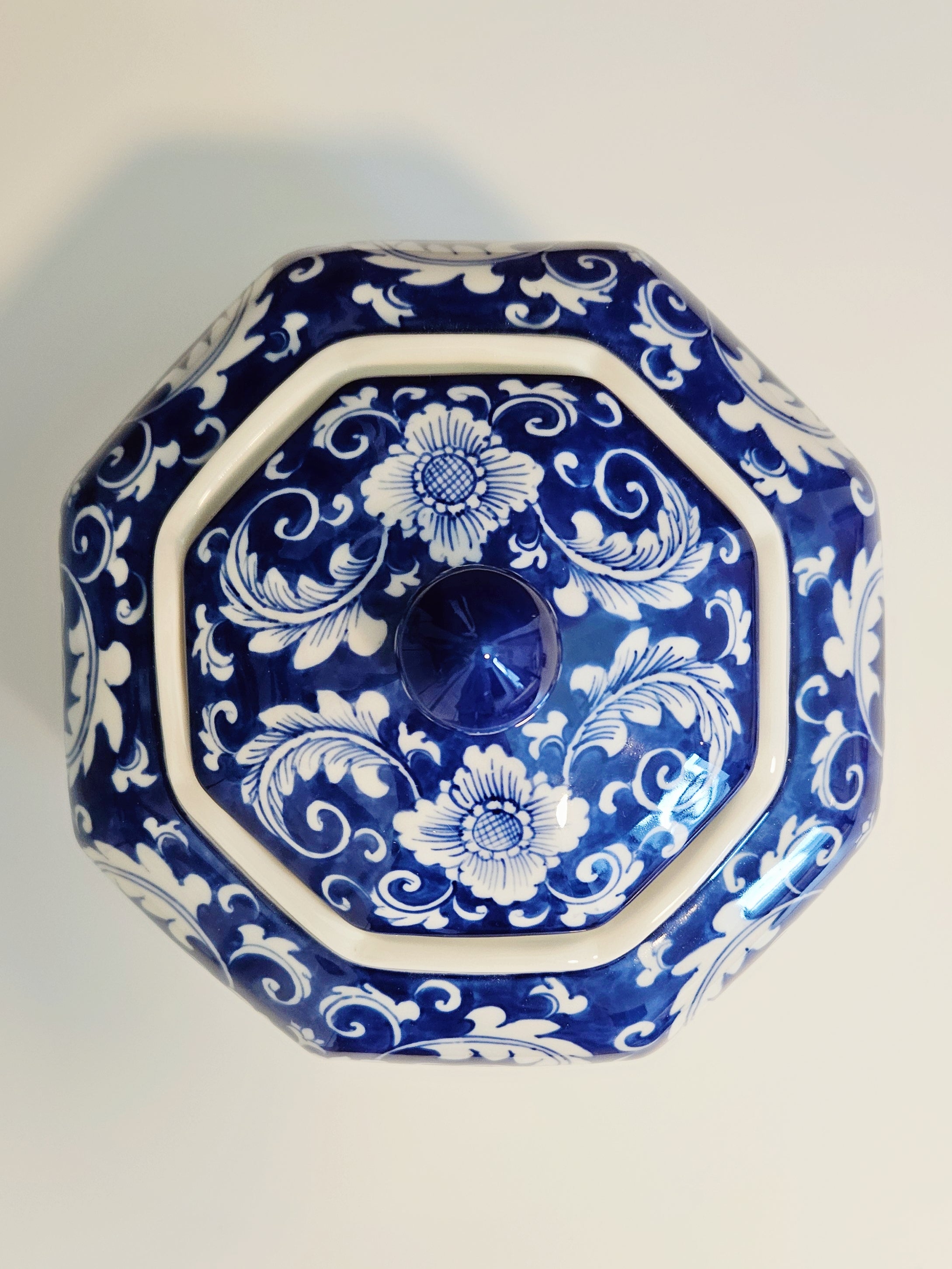 An octagonal ceramic lidded jar with a blue and white floral pattern, featuring a central blue knob, photographed from a top-down angle on a light background.