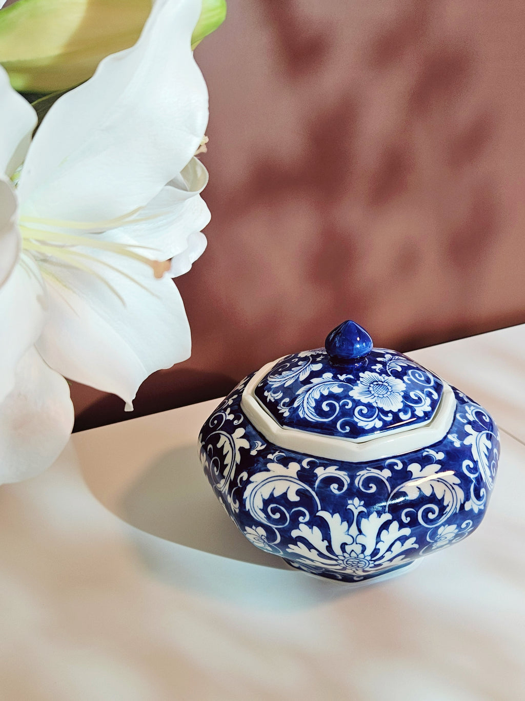 A chinoiserie blue and white porcelain canister with ornate floral scrollwork, styled on a light surface next to a large white flower against a warm brown background.