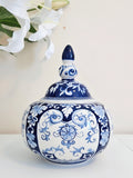 A chinoiserie blue and white porcelain lidded jar with intricate floral and scroll patterns, displayed on a white surface with white lilies in the background.