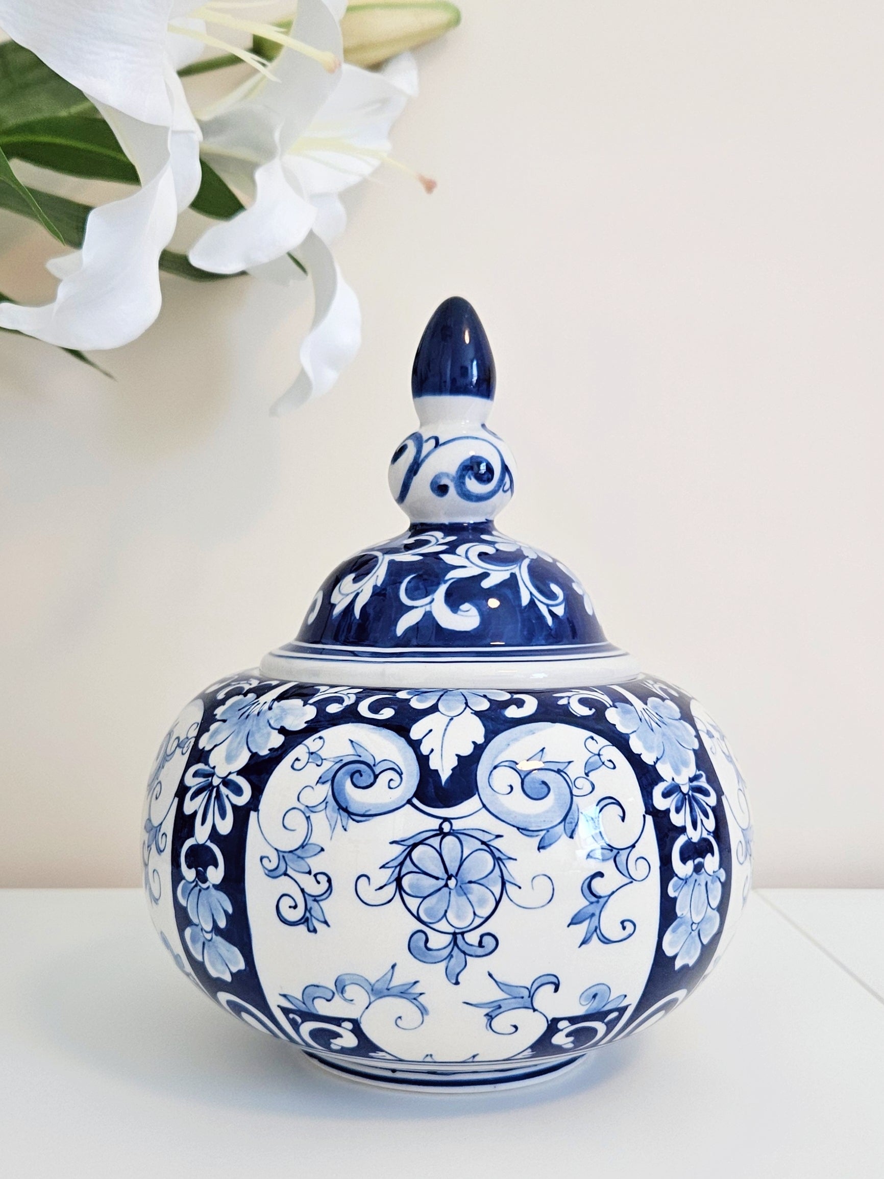A chinoiserie blue and white porcelain lidded jar with intricate floral and scroll patterns, displayed on a white surface with white lilies in the background.