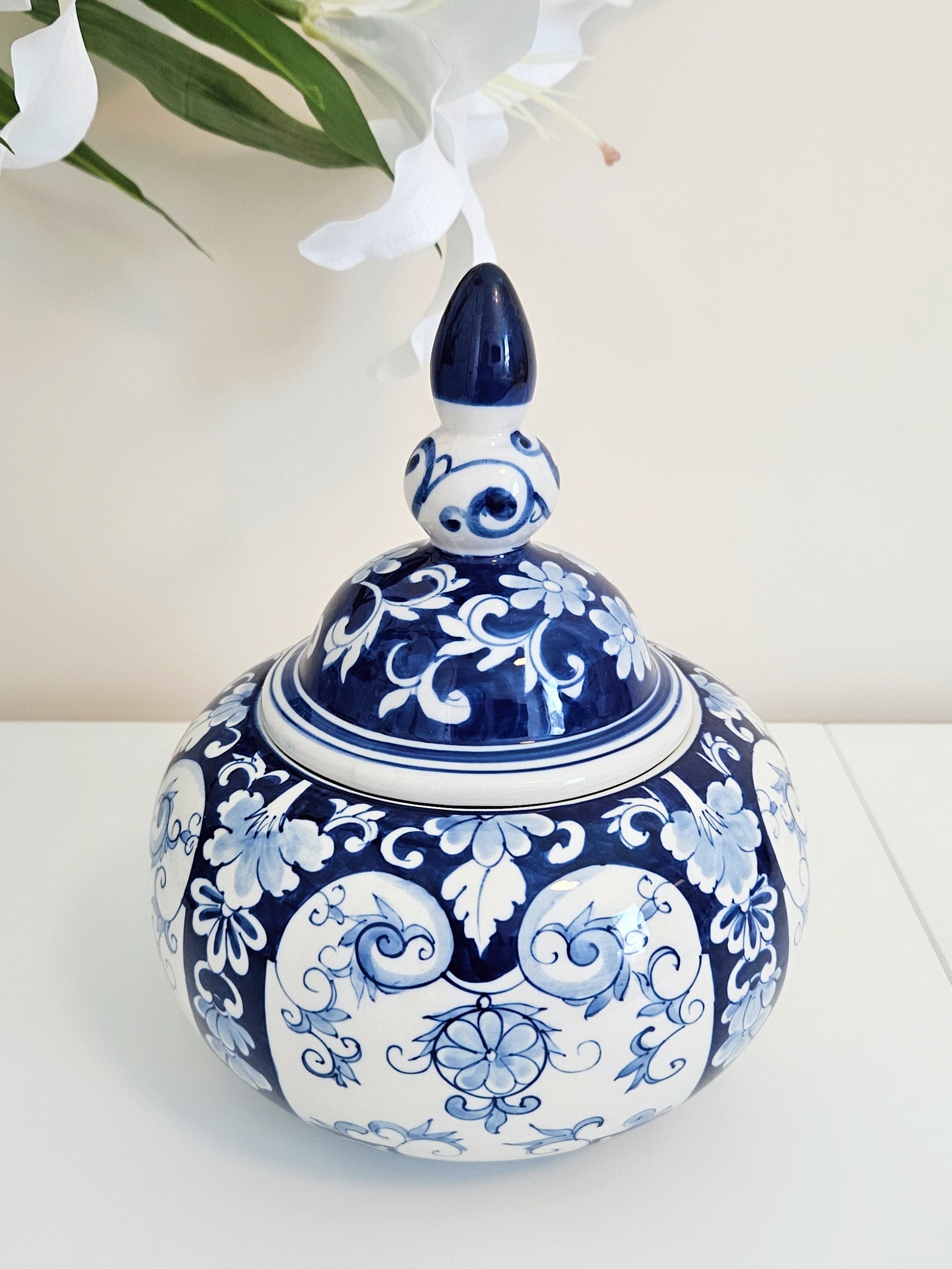 A round ceramic lidded jar or canister with a lid and pointed finial, decorated in a classic blue and white floral and scroll pattern, set on a white surface against a light beige background with white flowers in the upper left.