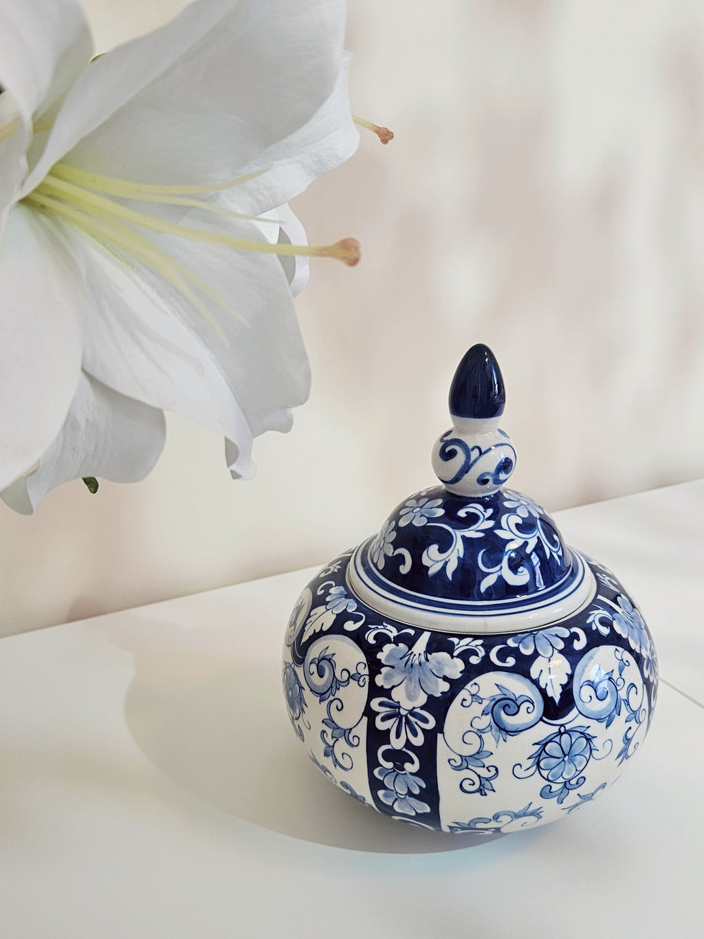 A chinoiserie blue and white porcelain lidded jar with intricate floral patterns, styled on a white surface next to a large white lily flower.