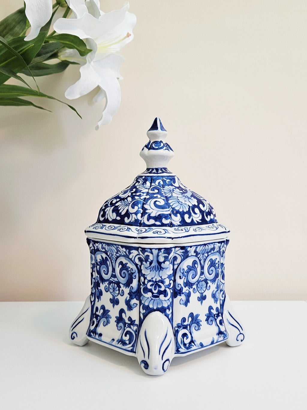 A chinoiserie blue and white porcelain lidded jar with ornate floral scrollwork, standing on a white surface against a light beige wall with white lilies in the upper left corner.