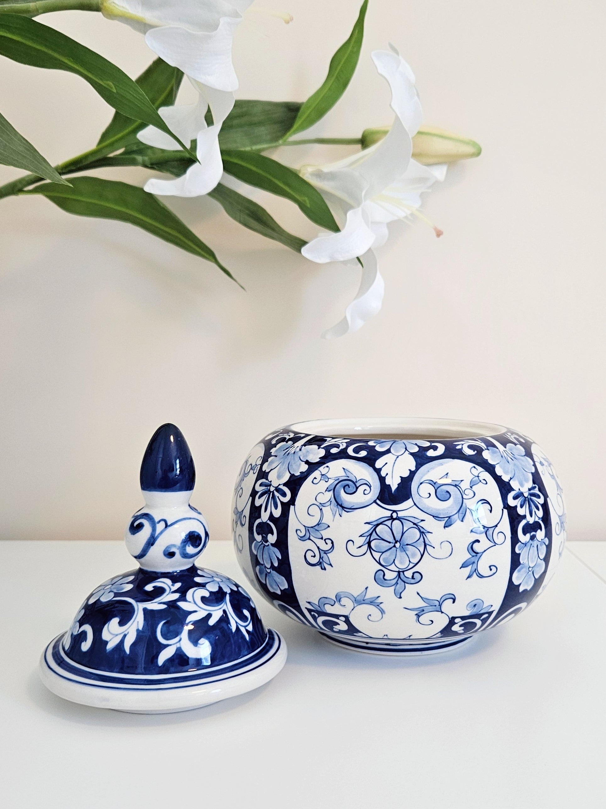 A blue and white porcelain lidded jar and its matching lid are displayed on a white surface with white lilies in the background.