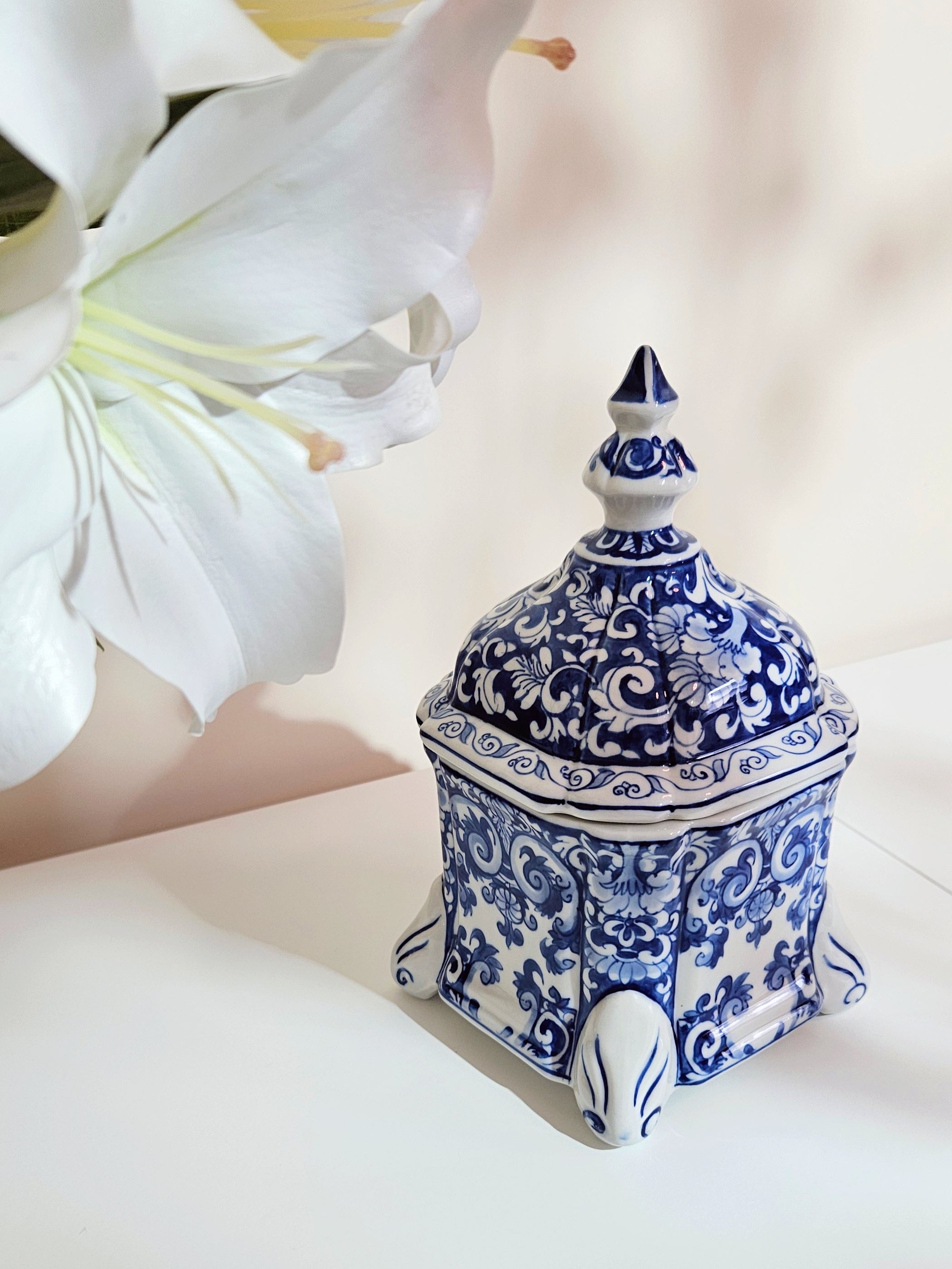 A small decorative chinoiserie lidded jar with a blue and white floral pattern, styled on a white surface next to a large white lily flower.