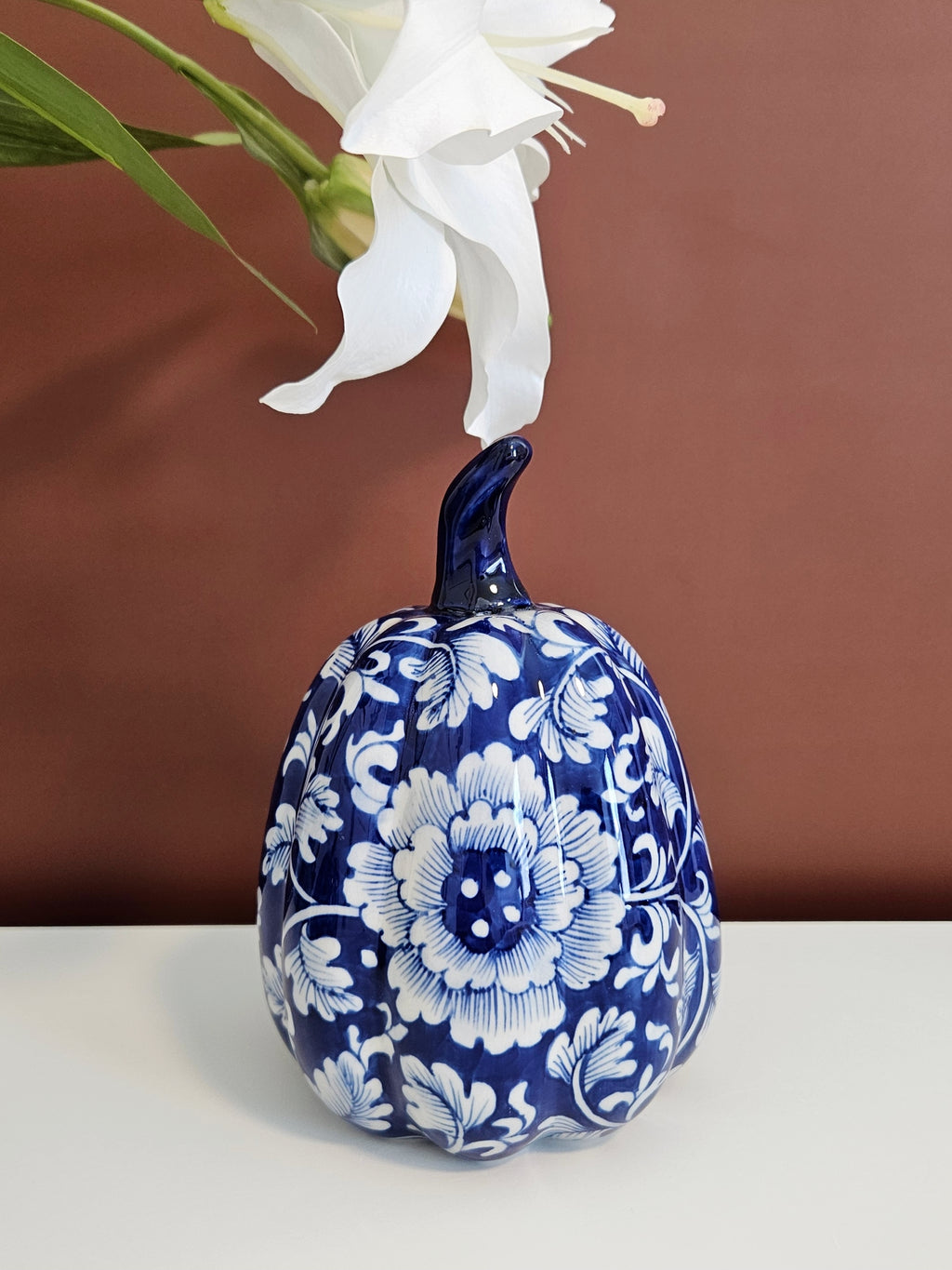 A decorative ceramic pumpkin figurine with a cobalt blue and white floral pattern, set against a solid brown background.