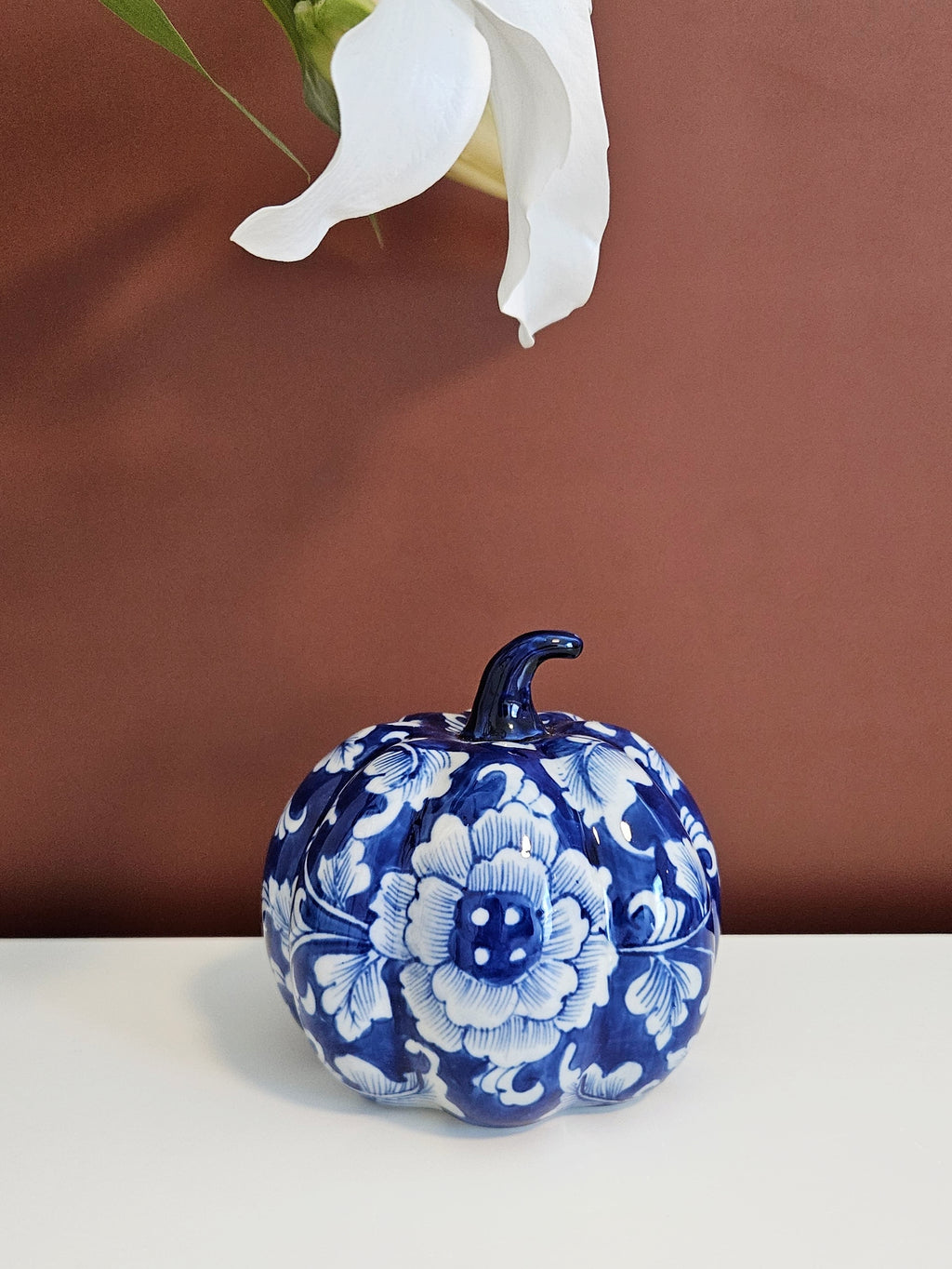 A decorative blue and white chinoiserie floral pumpkin-shaped figurine with a dark blue stem, placed on a white surface against a warm brown background with a white flower hanging above.