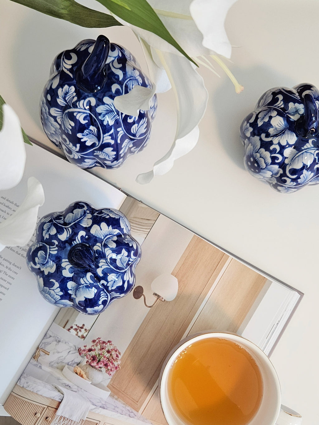 Three decorative ceramic pumpkins with a blue and white floral pattern are arranged on a white tabletop alongside an open book, a cup of tea, and white flowers.