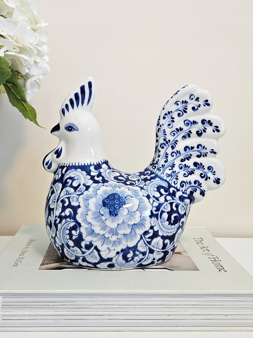 A decorative ceramic rooster figurine with a blue and white floral pattern, chinoiserie style, displayed on top of a stack of books with white flowers in the background.