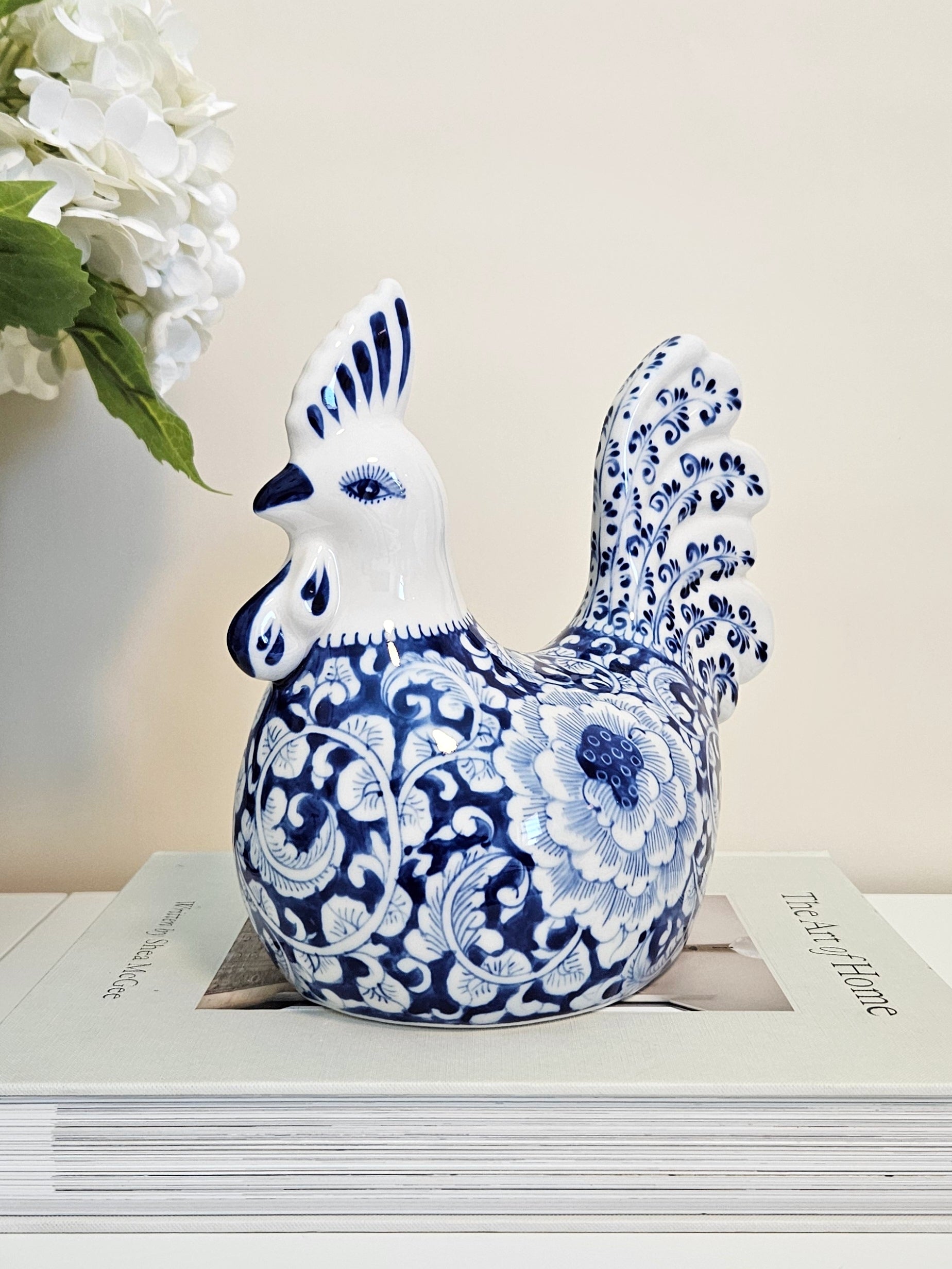 A hand-painted porcelain rooster figurine with a blue and white floral pattern, chinoiserie style,  displayed on a stack of books with white flowers in the background.