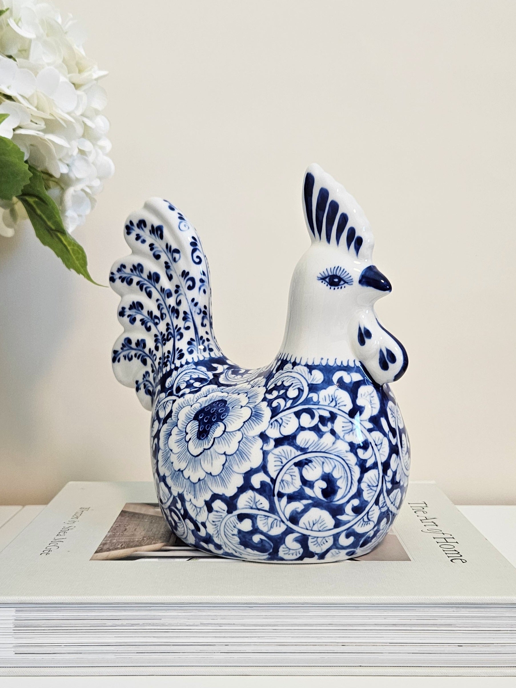 A decorative ceramic rooster figurine with a blue and white floral pattern, displayed on top of a stack of books against a light beige background.
