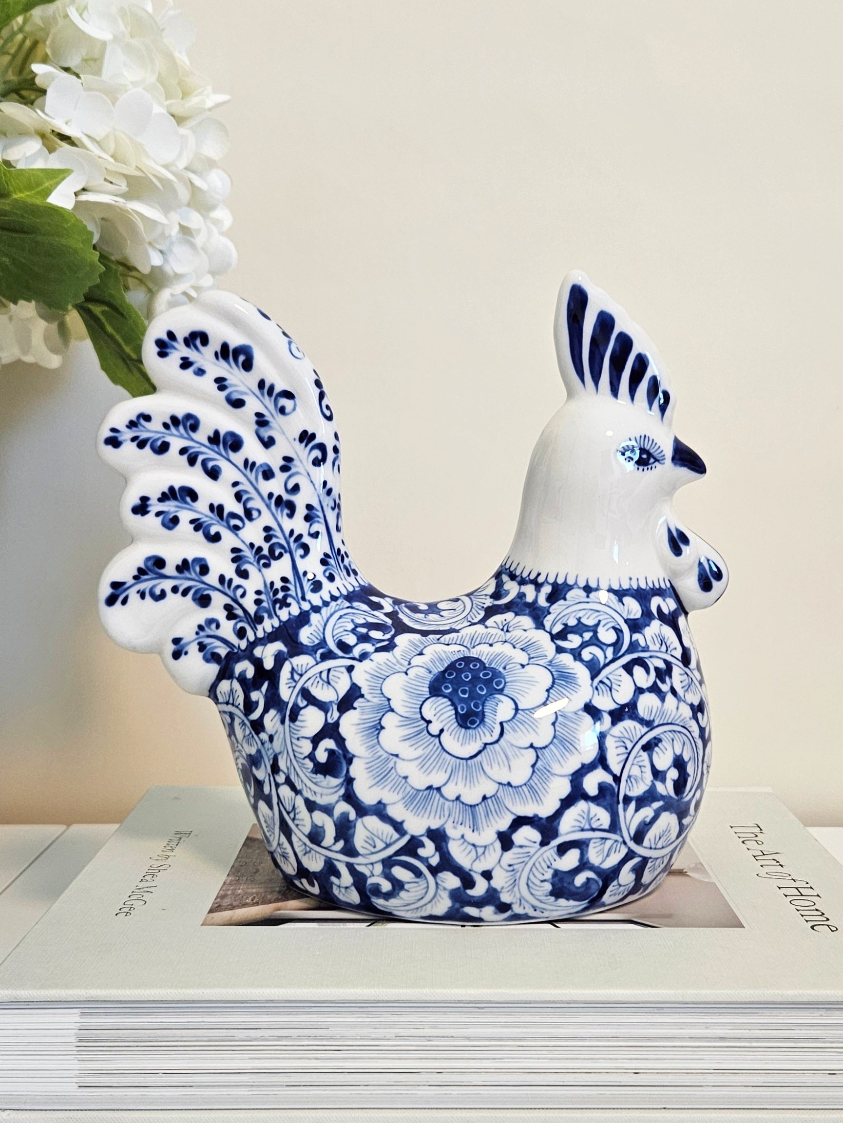 A decorative ceramic rooster figurine in a blue and white floral porcelain style, displayed on top of stacked books with white flowers in the background.