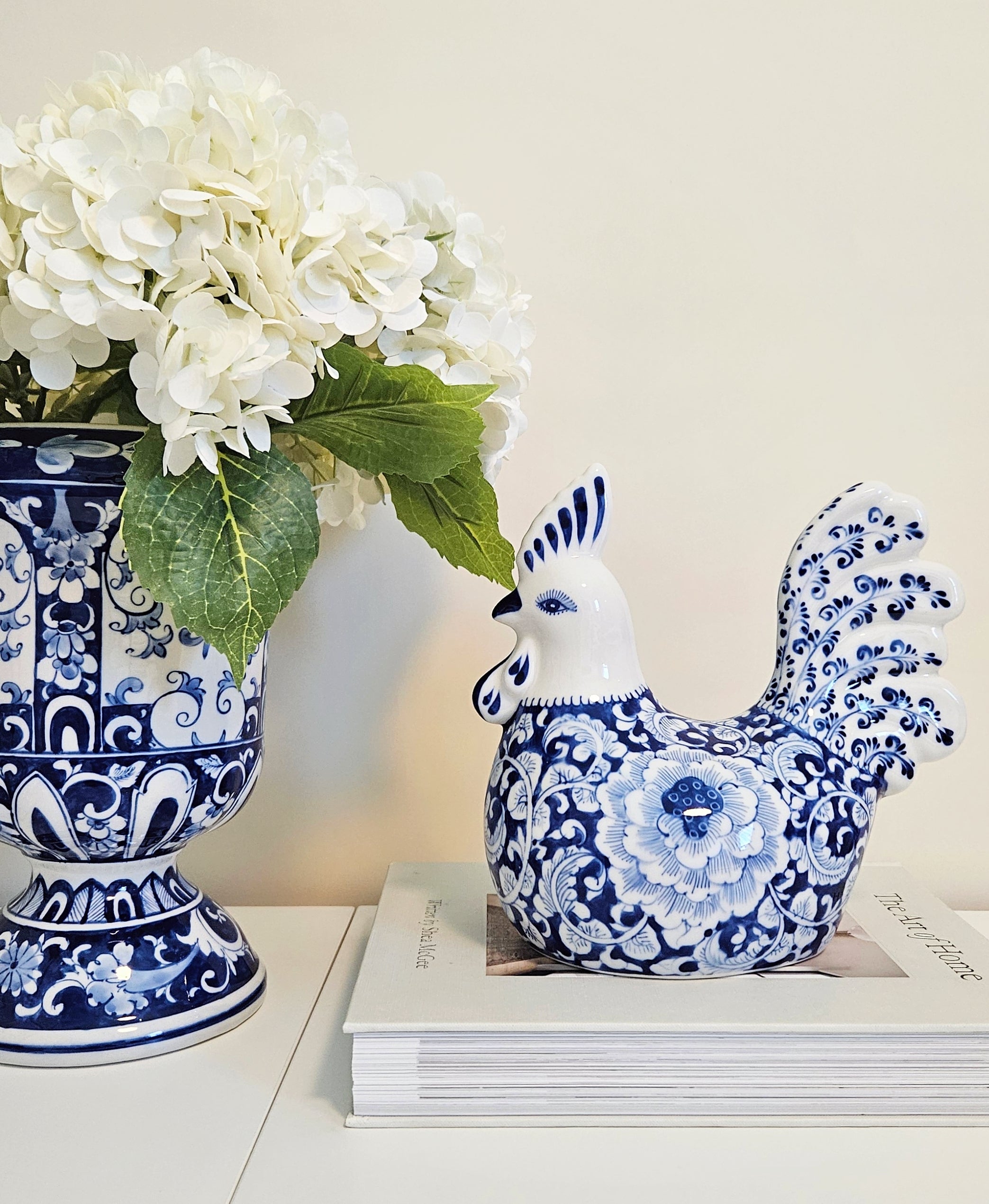 A blue and white porcelain rooster figurine displayed on a stack of books next to a matching blue and white floral vase with white flowers against a warm beige background.