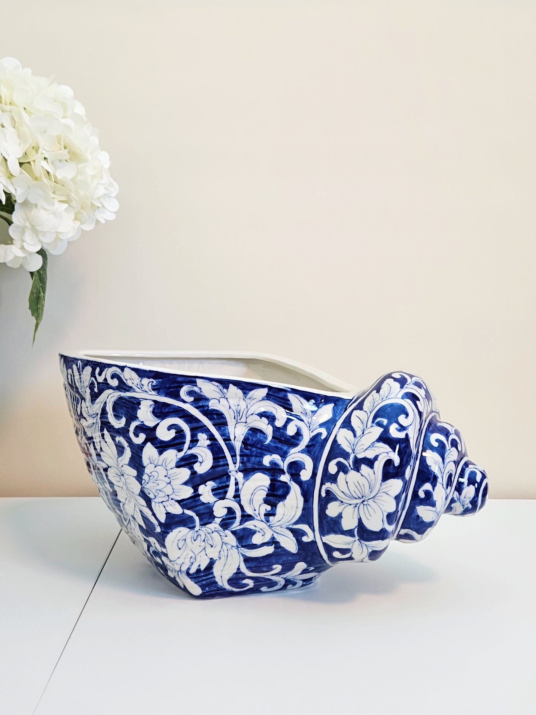A ceramic planter shaped like a seashell, decorated with a blue and white floral pattern, sitting on a white surface against a beige wall with white flowers in the background.