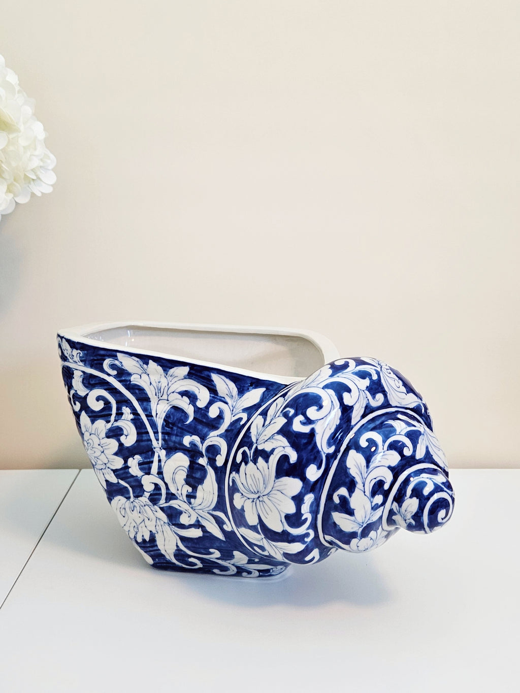 A sculptural ceramic planter shaped like a spiral shell, featuring a glossy blue and white floral baroque pattern, set against a neutral cream background.