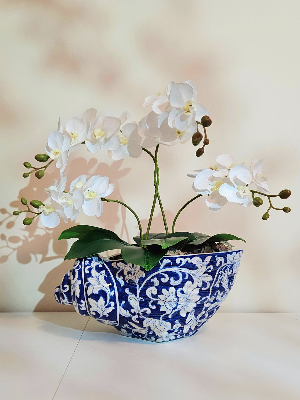 White artificial orchid flowers with green leaves arranged in a blue and white patterned ceramic bowl planter, set against a warm beige background with soft leaf shadows.