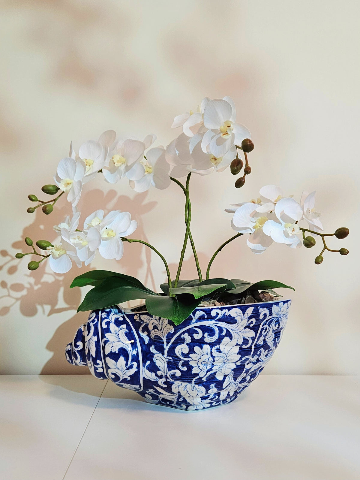 White artificial orchid flowers with green leaves arranged in a blue and white patterned ceramic bowl planter, set against a warm beige background with soft leaf shadows.