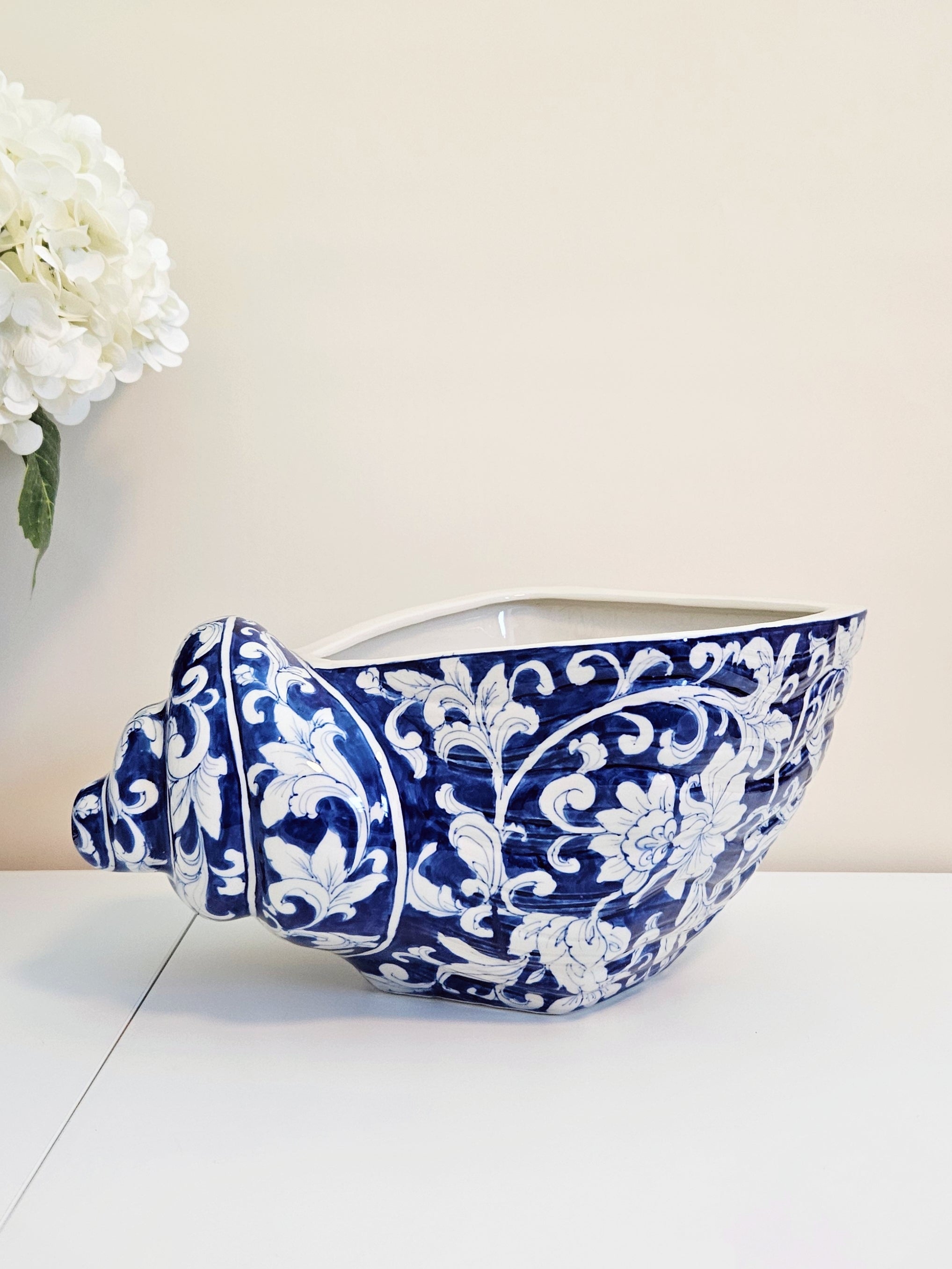 A ceramic planter shaped like a seashell, featuring a deep cobalt blue and white floral pattern, displayed on a white surface against a light beige wall with white flowers in the background.