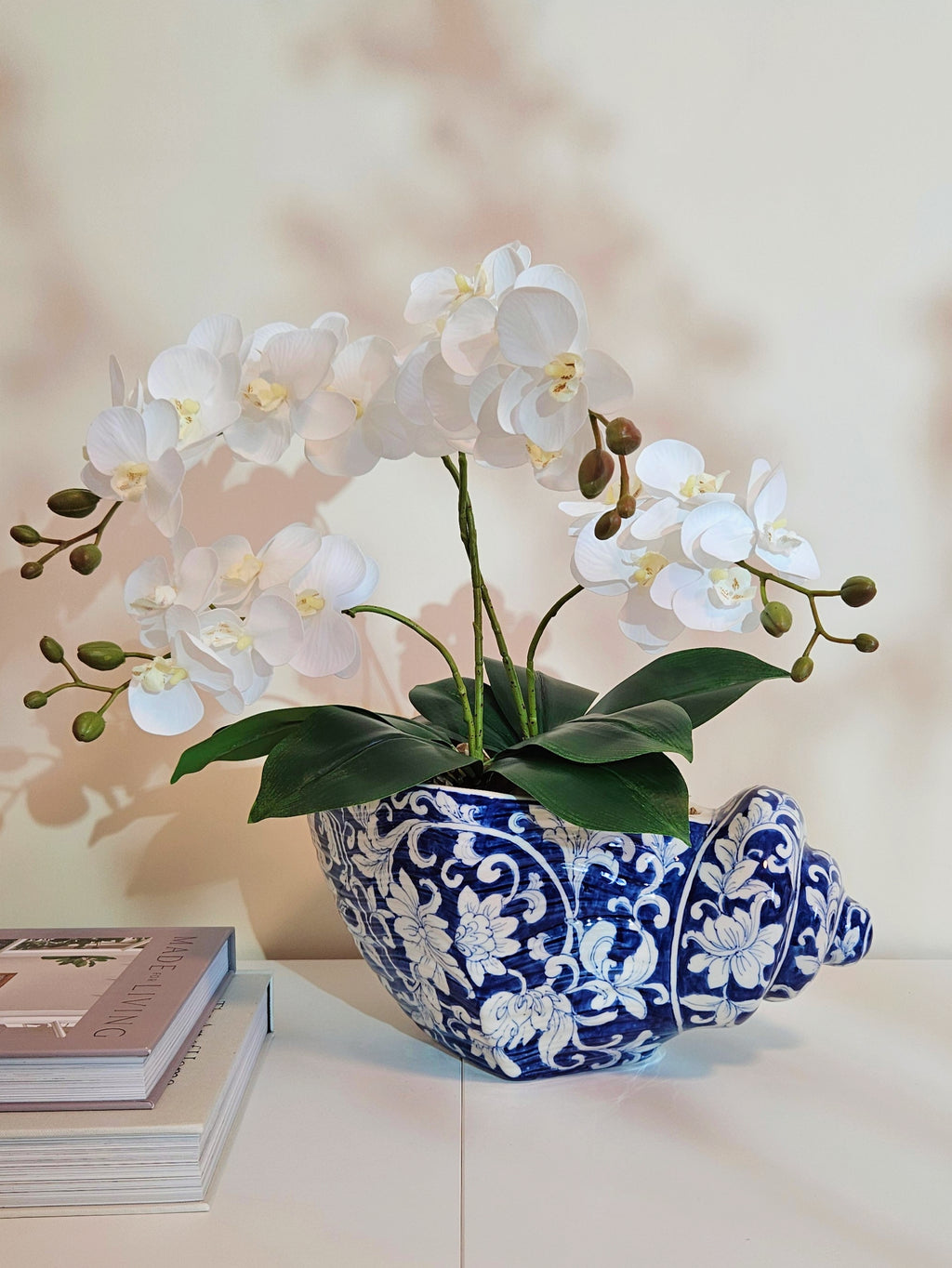 White artificial orchid flowers arranged in a blue and white porcelain shell-shaped planter on a white tabletop, with stacked books partially visible on the left against a softly lit beige wall.