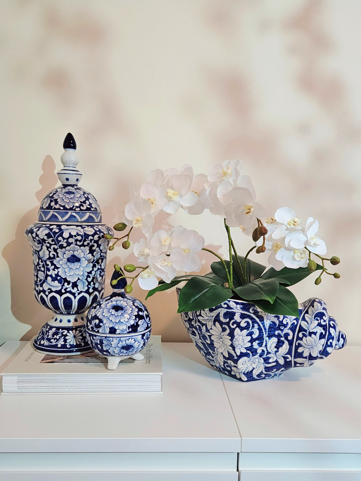 Blue and white porcelain decorative pieces on a white table, including a lidded jar, a small lidded box, and a sculptural planter shaped like a seashell holding white flowers, set against a softly lit beige wall.