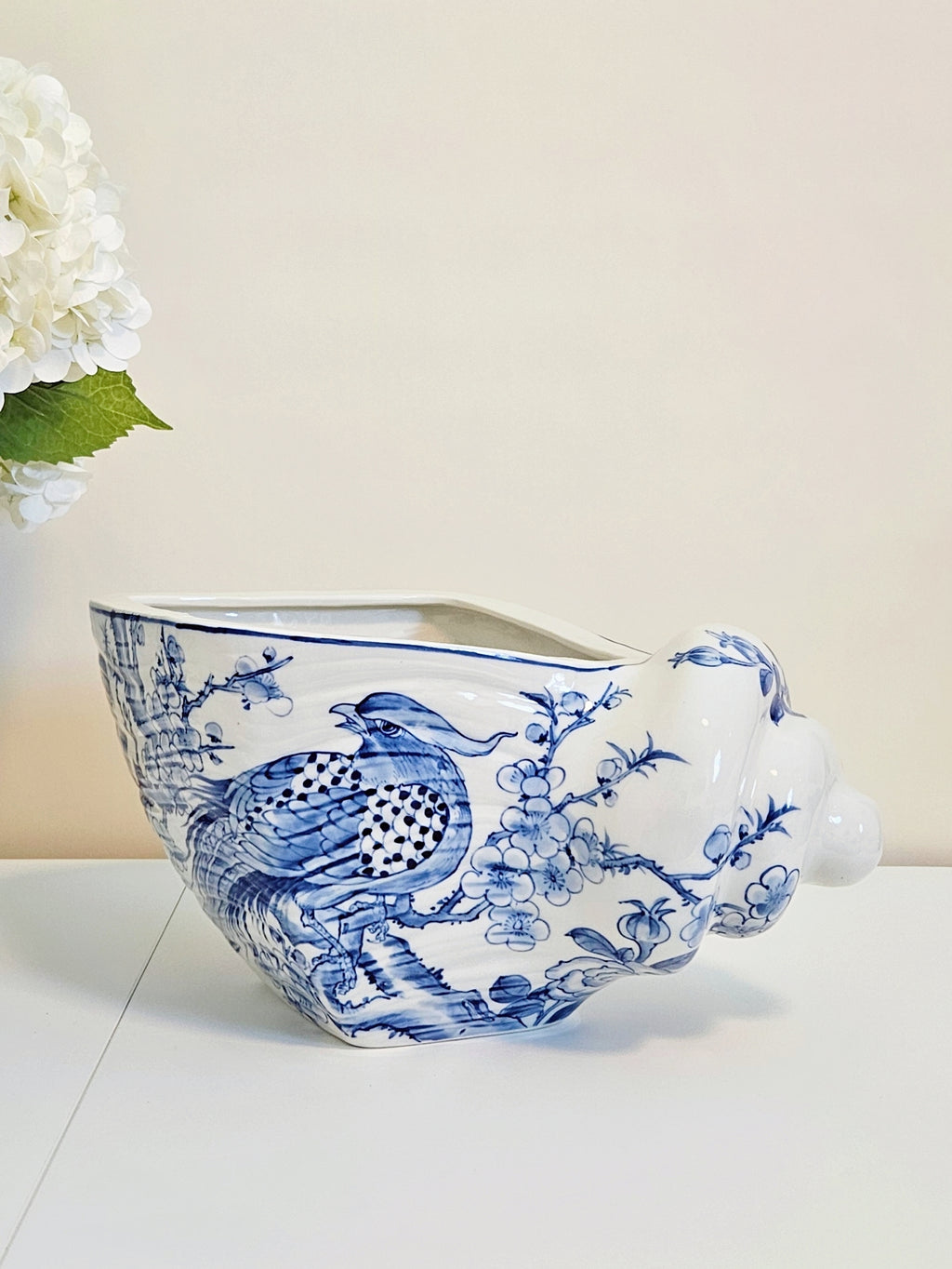 Hand-painted blue and white chinoiserie porcelain shell planter pot with pheasants and cherry blossoms motif, placing on a white surface with beige background behind it