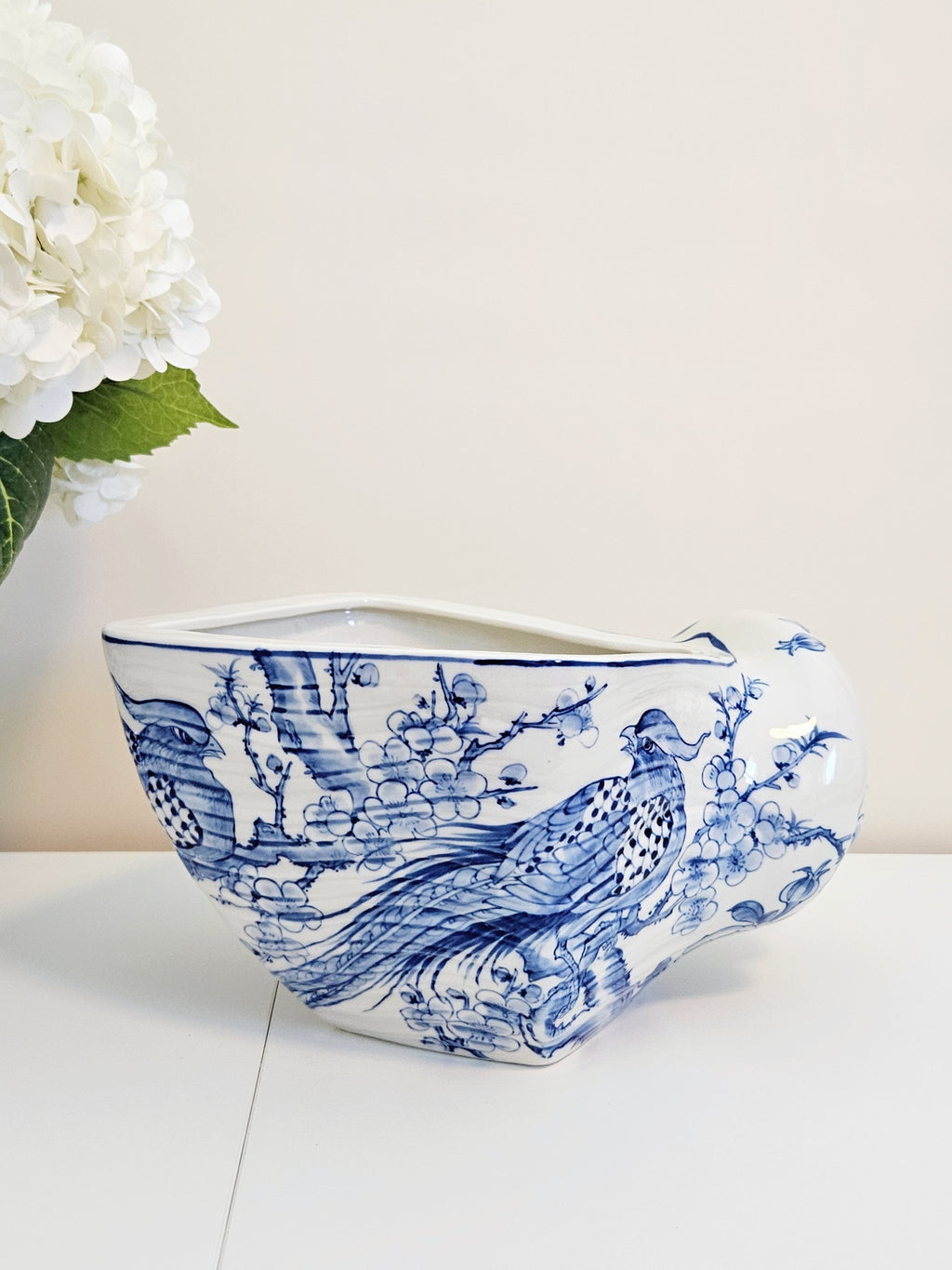 A white ceramic planter with a blue hand-painted traditional Asian style pheasants and floral design, styled on a white surface with a soft beige background and white flowers in the corner.