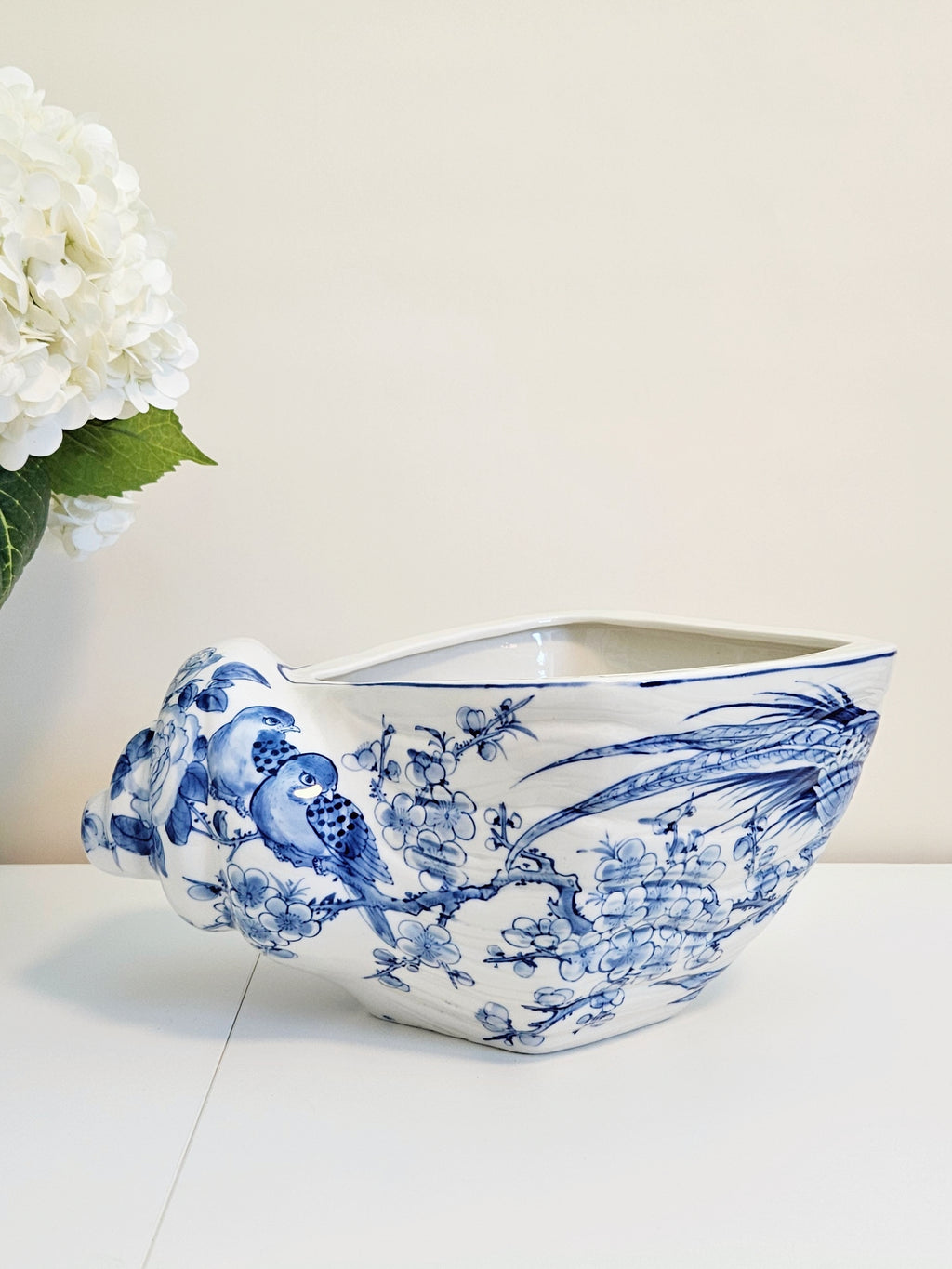 A white chinoiserie ceramic planter shaped like a shell, decorated with blue and white hand-painted bird and floral motifs, set on a white surface against a light beige background with white flowers on the left.