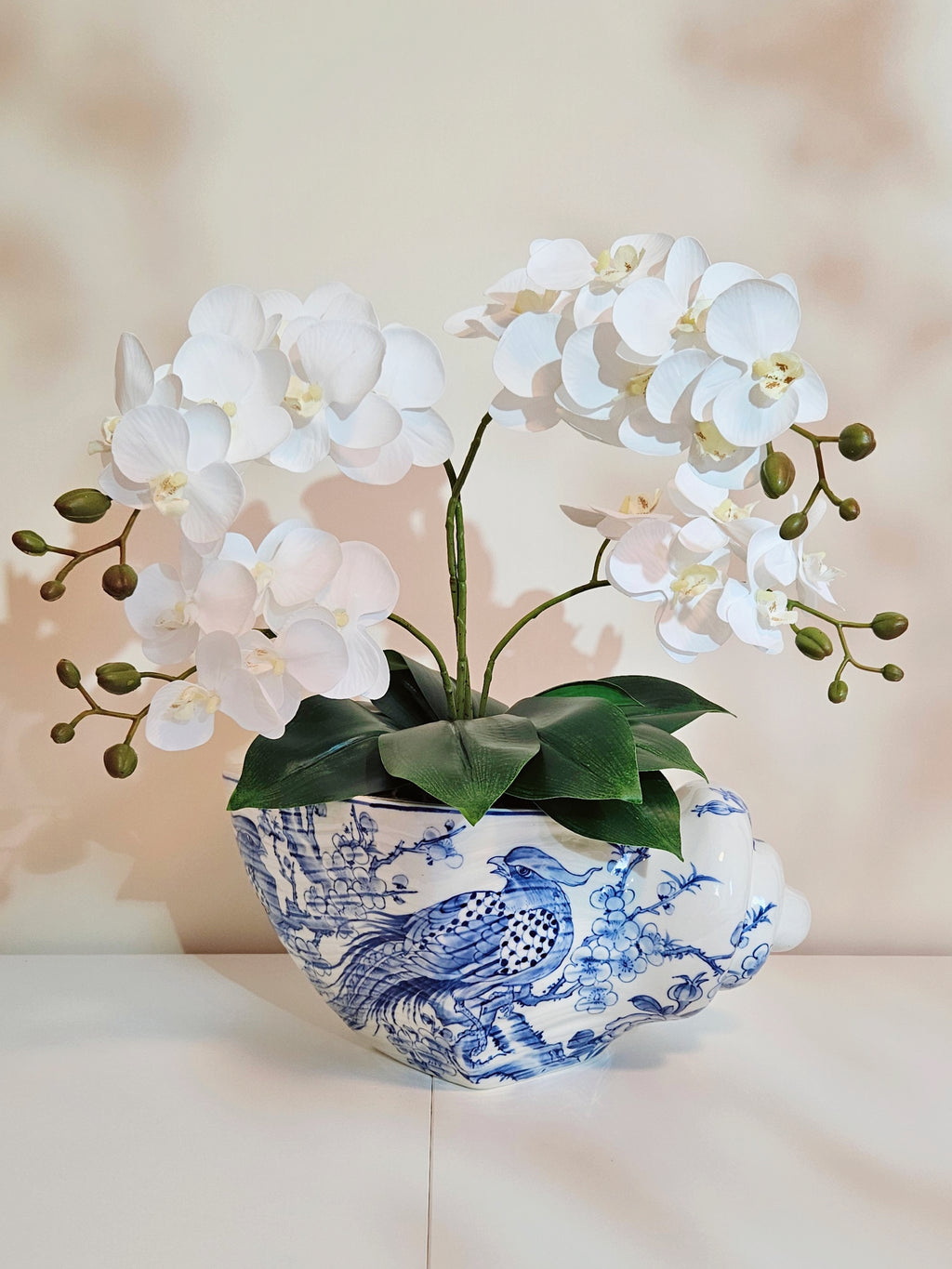 White artificial orchid flowers with green leaves arranged in a blue and white porcelain shell planter, set against a soft beige background.