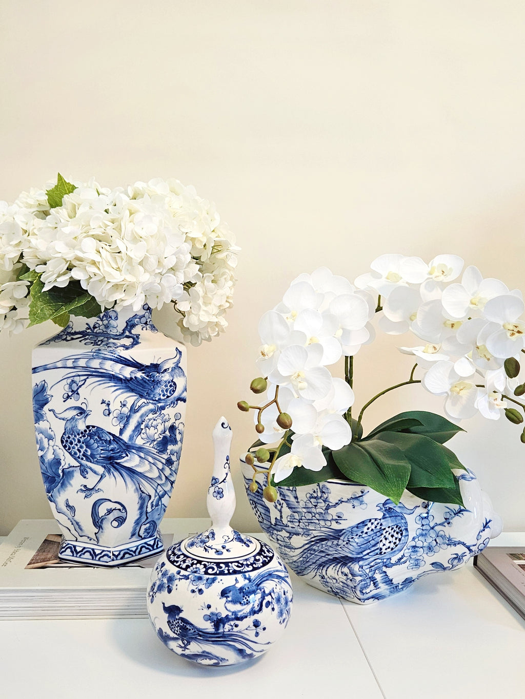 Three blue and white porcelain decorative pieces displayed on a white surface against a beige wall: a tall vase with white flowers, a matching planter with white orchids, and a small lidded jar with a decorative handle.
