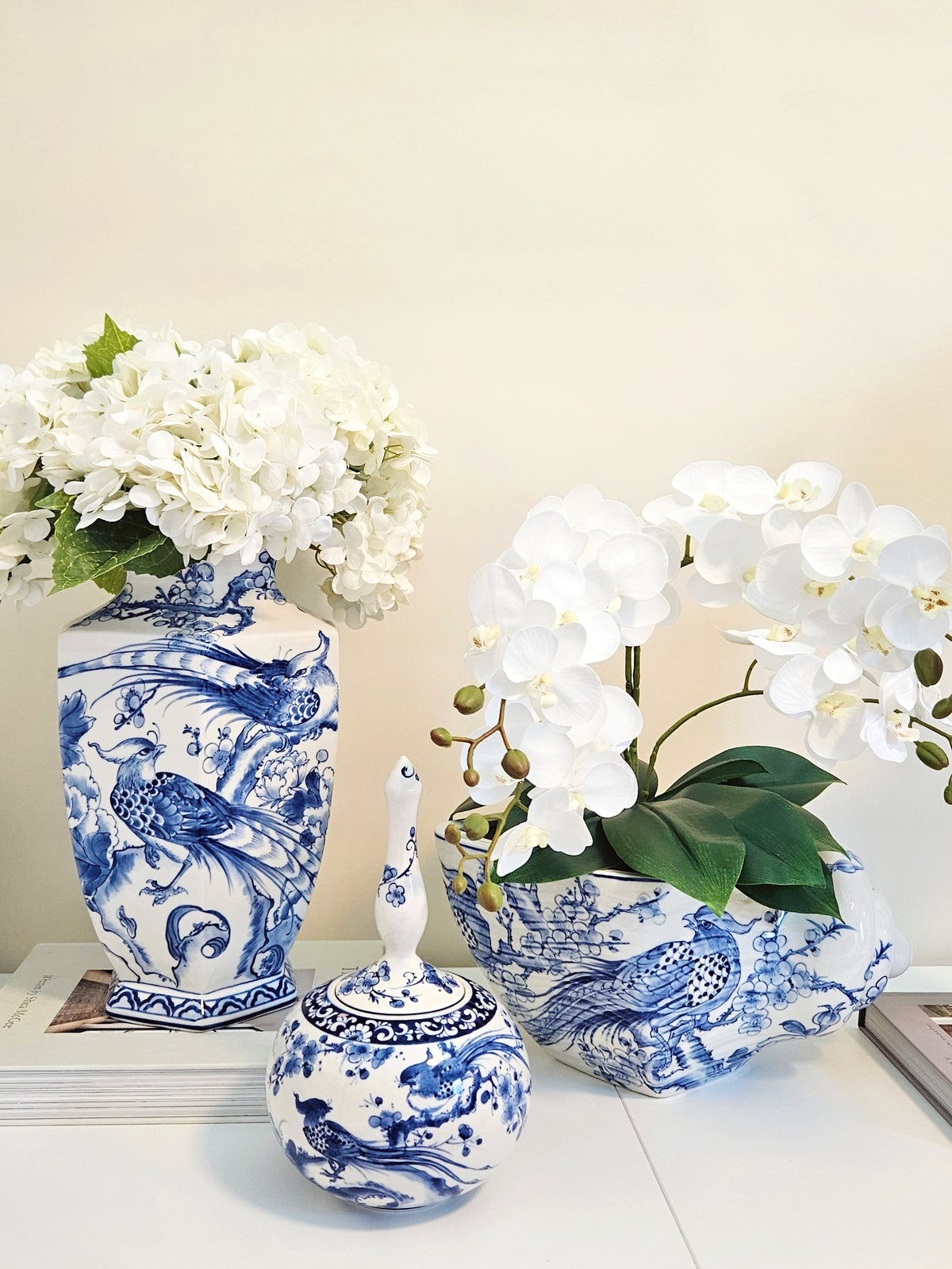 Three blue and white porcelain decorative pieces displayed on a white surface against a beige wall: a tall vase with white flowers, a matching planter with white orchids, and a small lidded jar with a decorative handle.