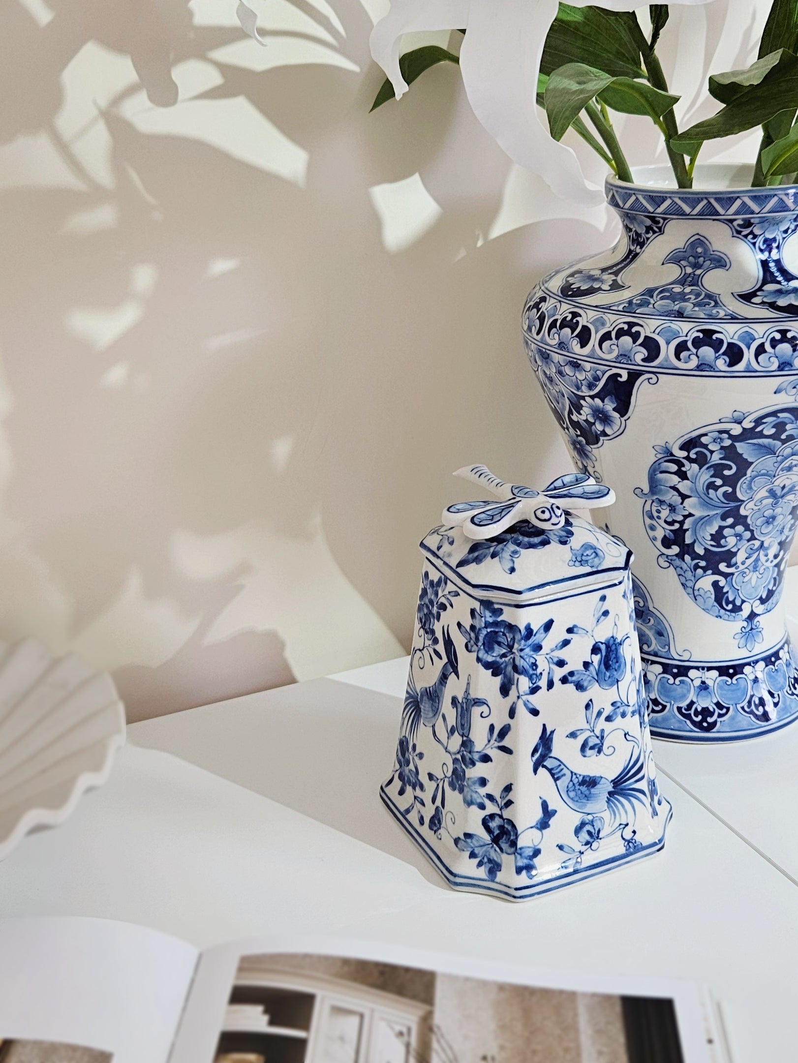 Blue and white porcelain decorative pieces, including a small lidded canister with a floral bird pattern and bow on top, and a larger vase with matching blue floral motifs, styled on a white surface with soft shadow overlays.