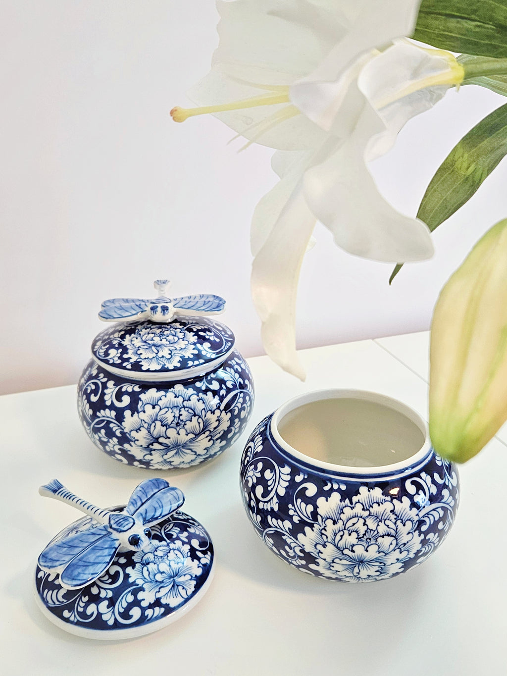A chinoiserie blue and white floral ceramic jars with dragonfly lids, styled with white lilies on a white surface against a white background.