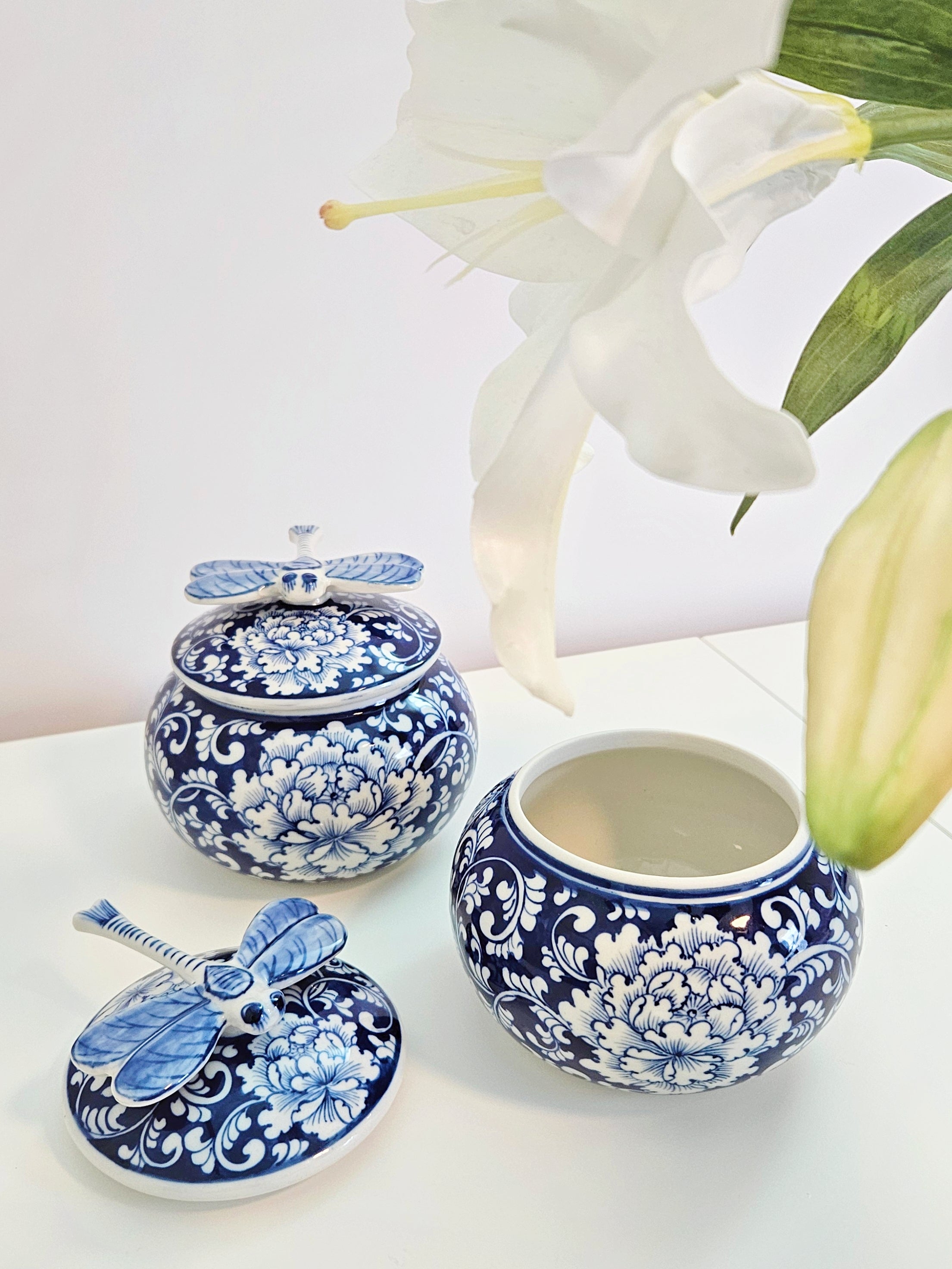 A chinoiserie blue and white floral ceramic jars with dragonfly lids, styled with white lilies on a white surface against a white background.
